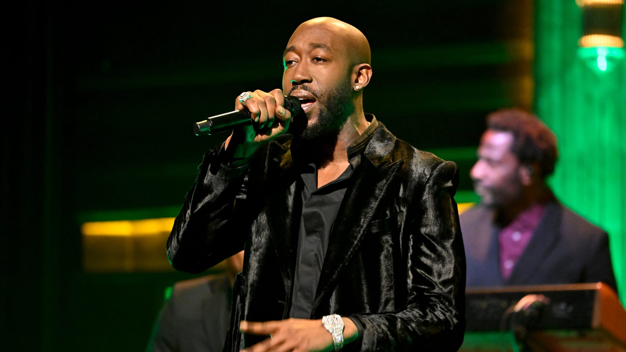 Man in velvet jacket singing into microphone on stage with band member in background