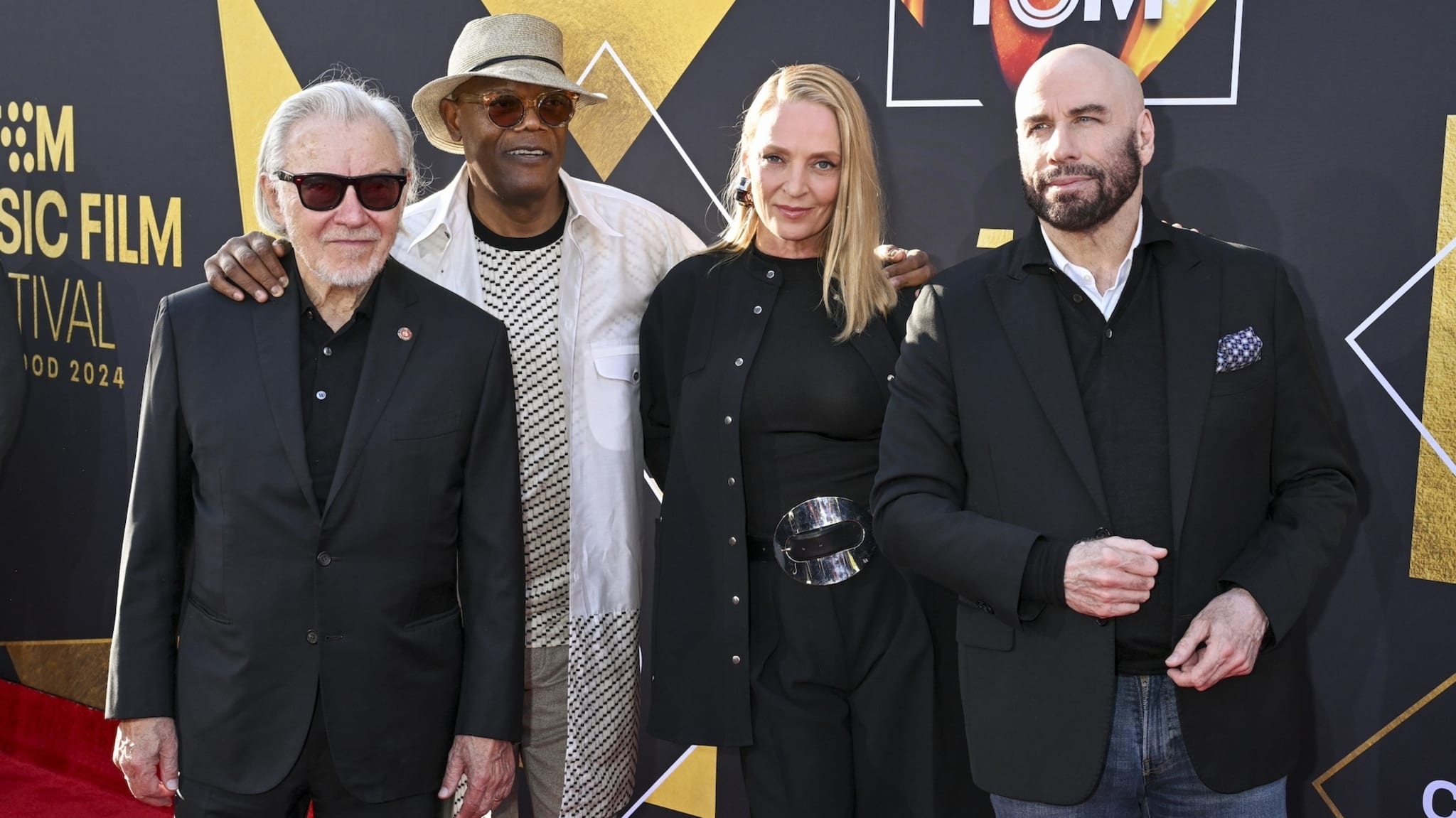 Four people posing together at an event, with two men in suits, a woman in a dress and belt, and a man in a hat and jacket