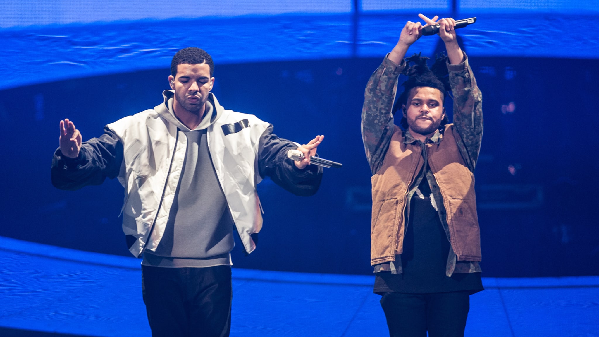 Two musicians performing onstage, one gesturing while the other holds a microphone up