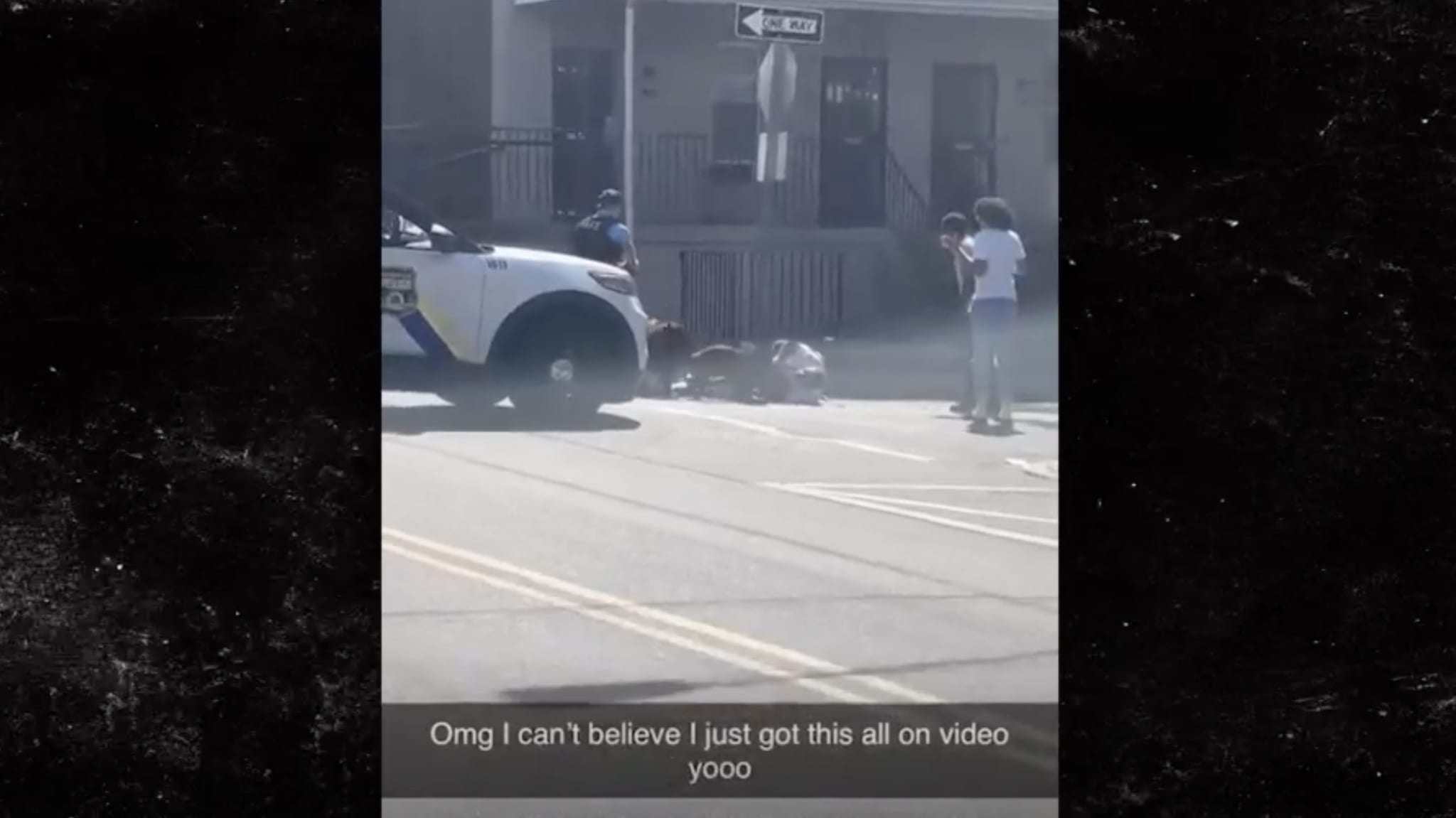 A blurred action scene with a person filming, another standing by, and a figure on the ground near a car