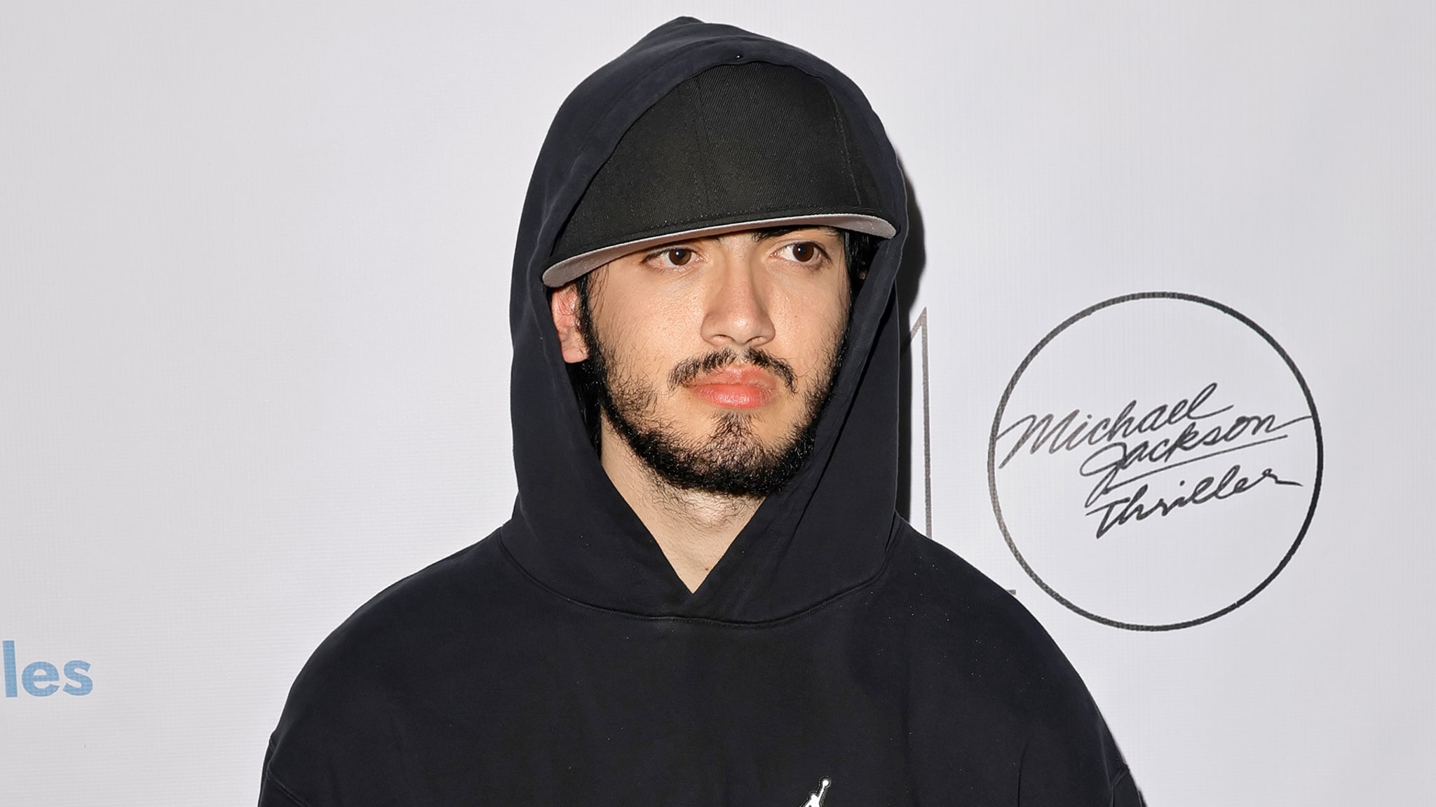 Man in a hooded black outfit standing in front of a promotional backdrop