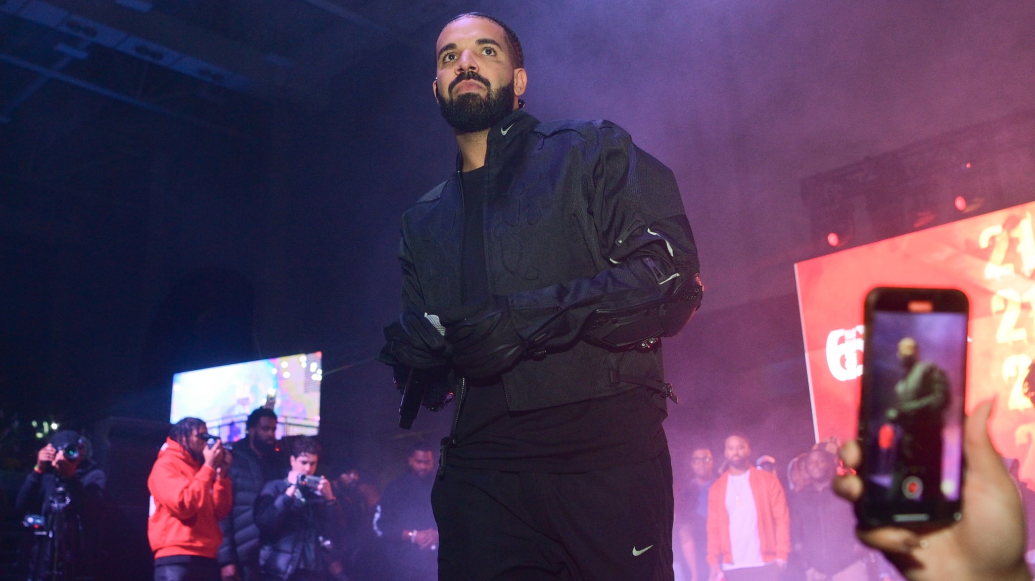 Drake performing on stage, wearing a black jacket, with fans capturing the moment on their phones