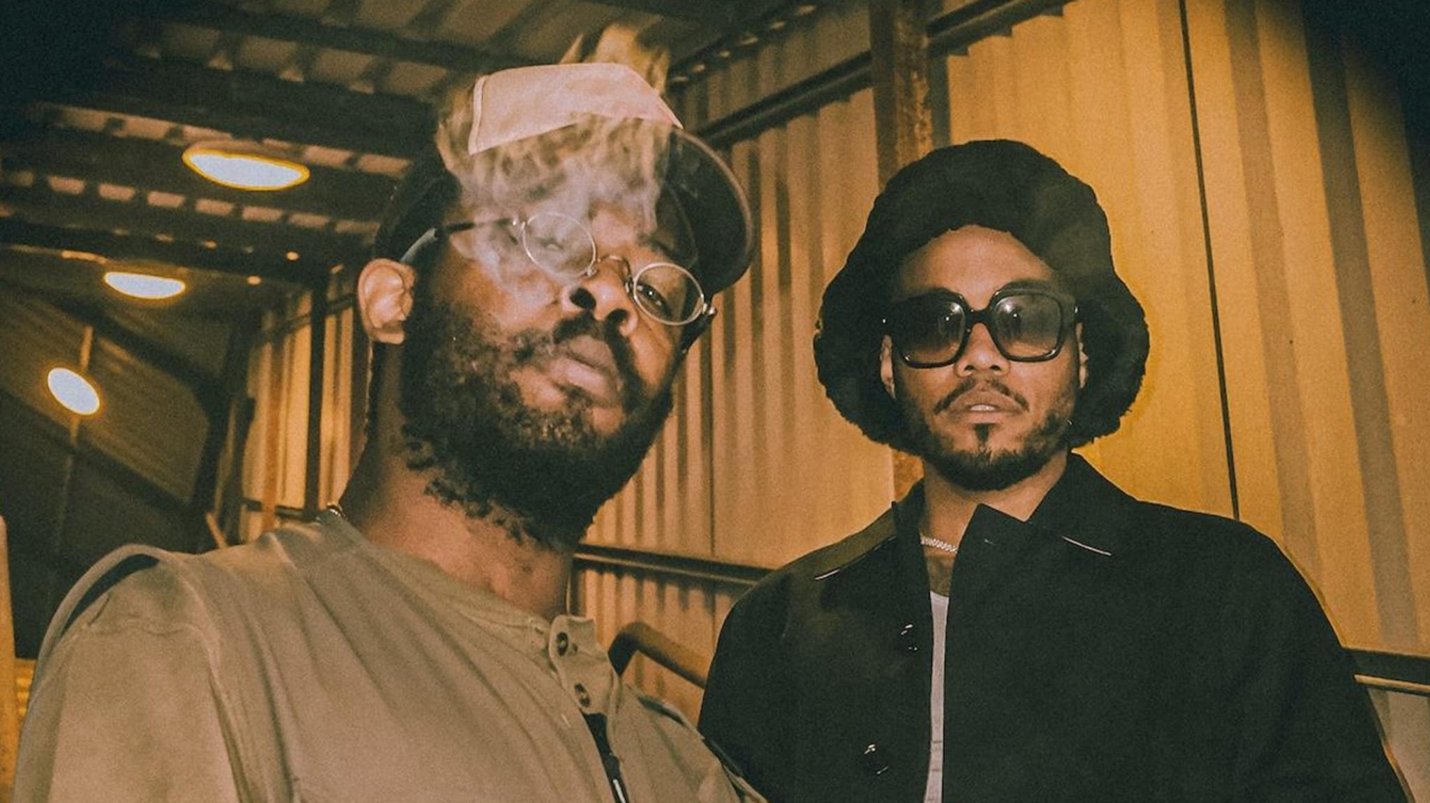 Two men posing for a photo, one exhaling smoke, both wearing hats and casual jackets