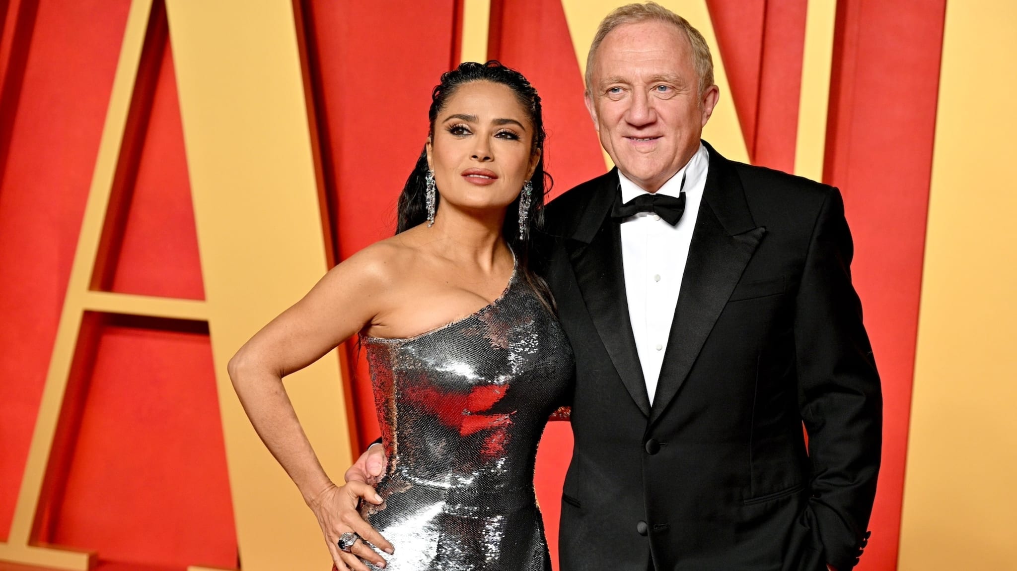 Two celebrities posing; the woman in a metallic gown, the man in a classic tuxedo, at a gala event