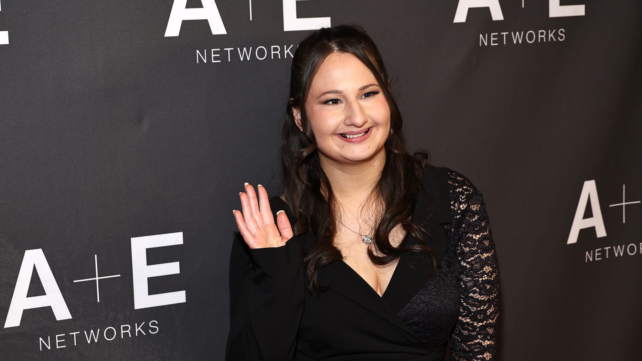 Woman on media backdrop smiles and waves, in black sleeveless dress with lace detail