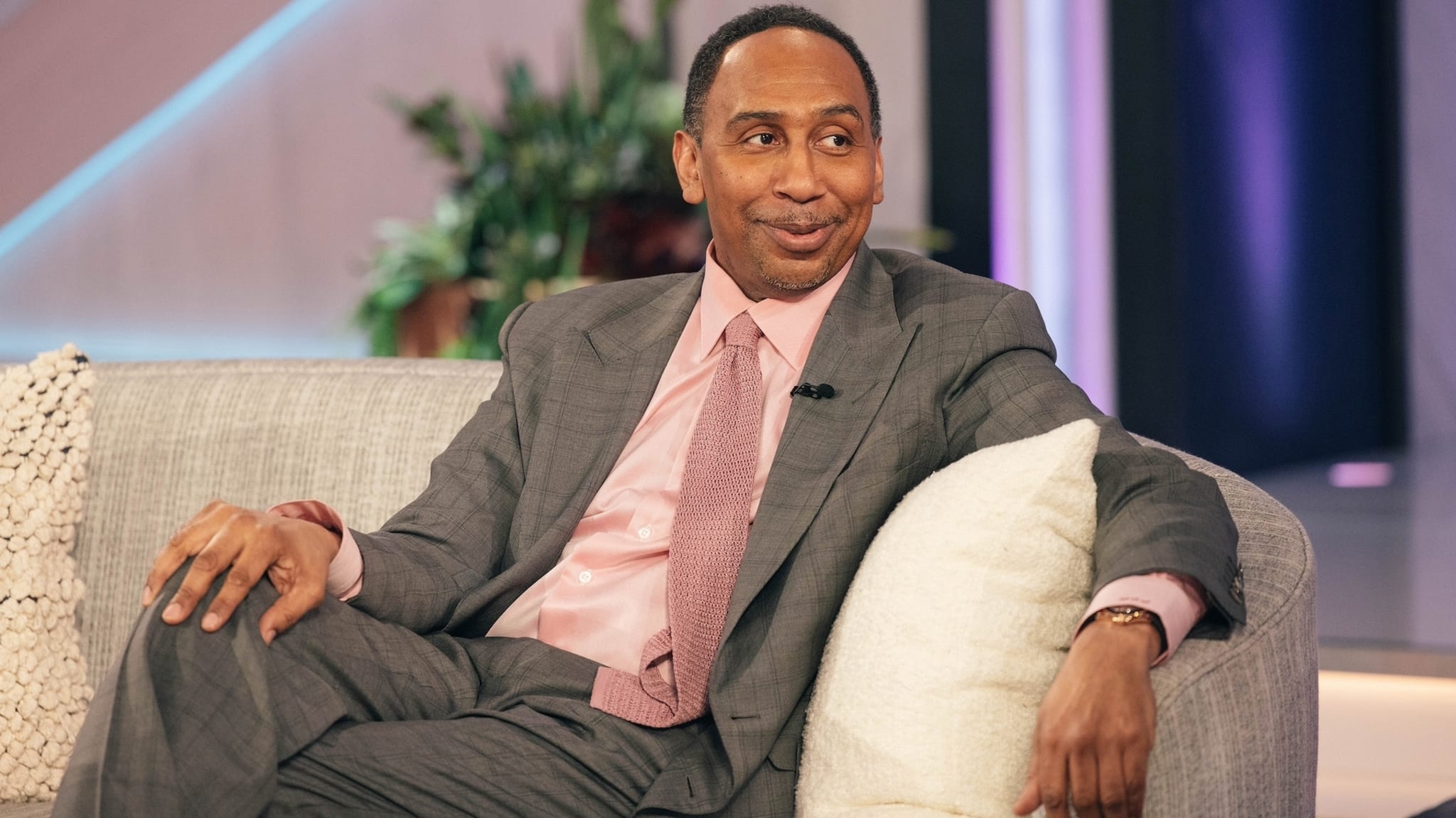 TV show guest wearing a gray suit and pink tie sitting on a couch, smiling