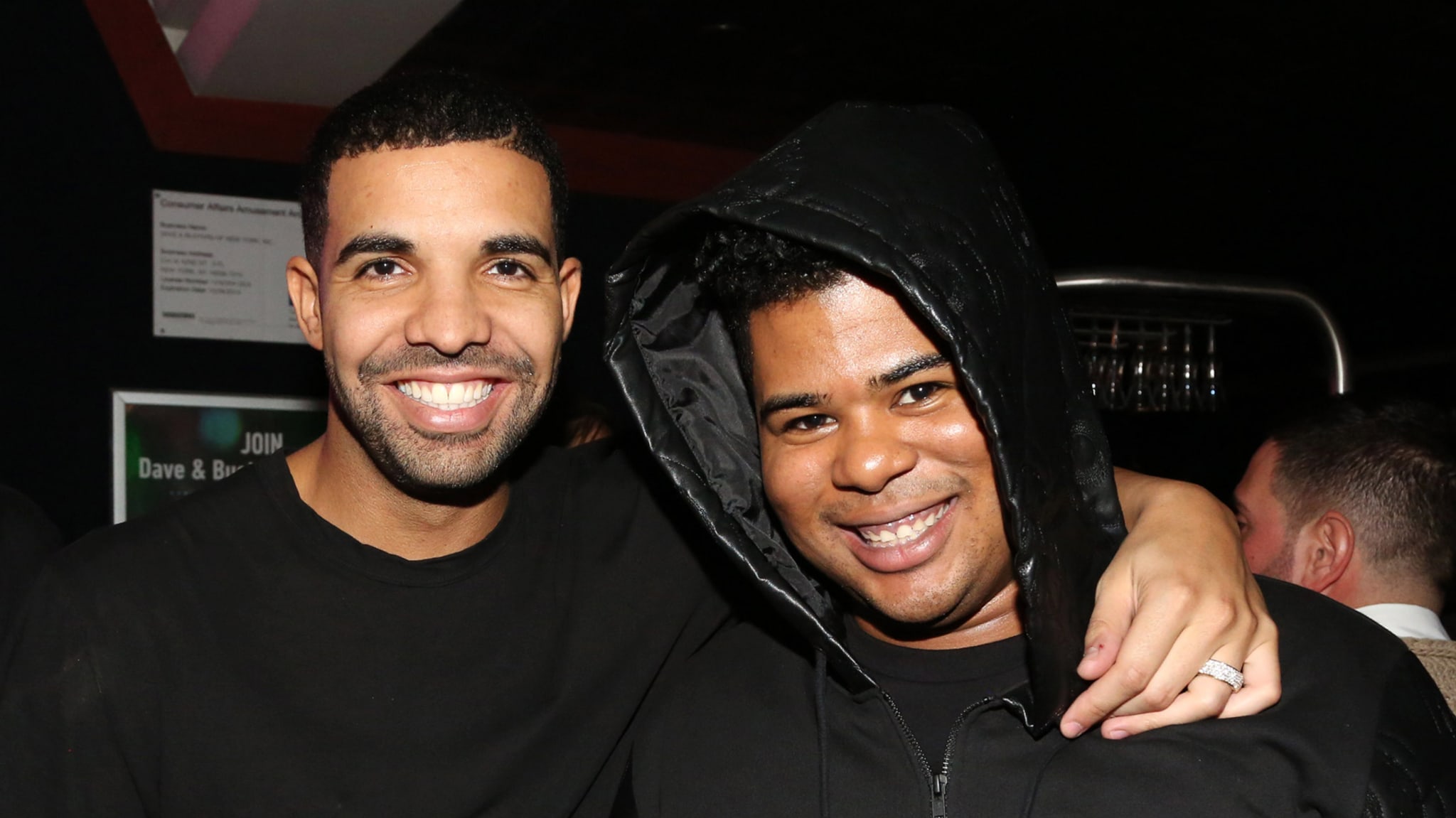 Two men smiling with their arms around each other, one wearing a black hoodie