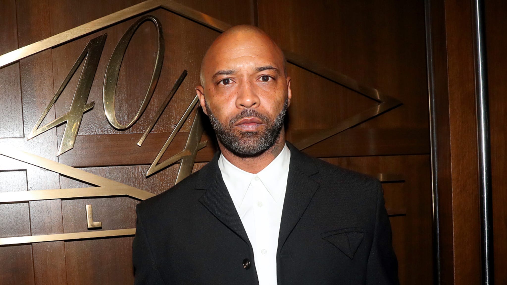 Joe Budden in a black suit with a lapel pin, standing before a wooden backdrop with '40/40 Club' etched in it