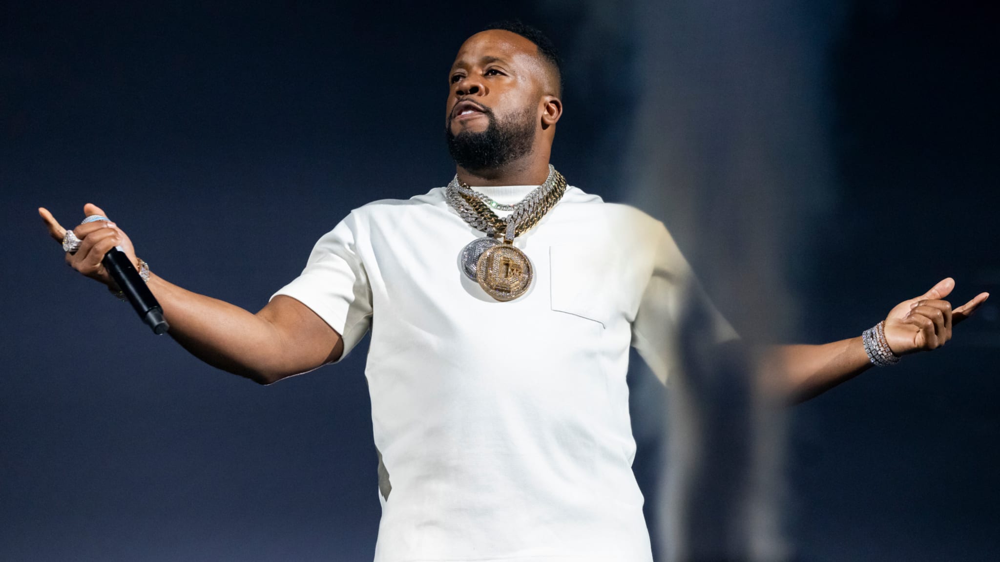 Yo Gotti on stage performing, wearing a white t-shirt and gold chain necklace