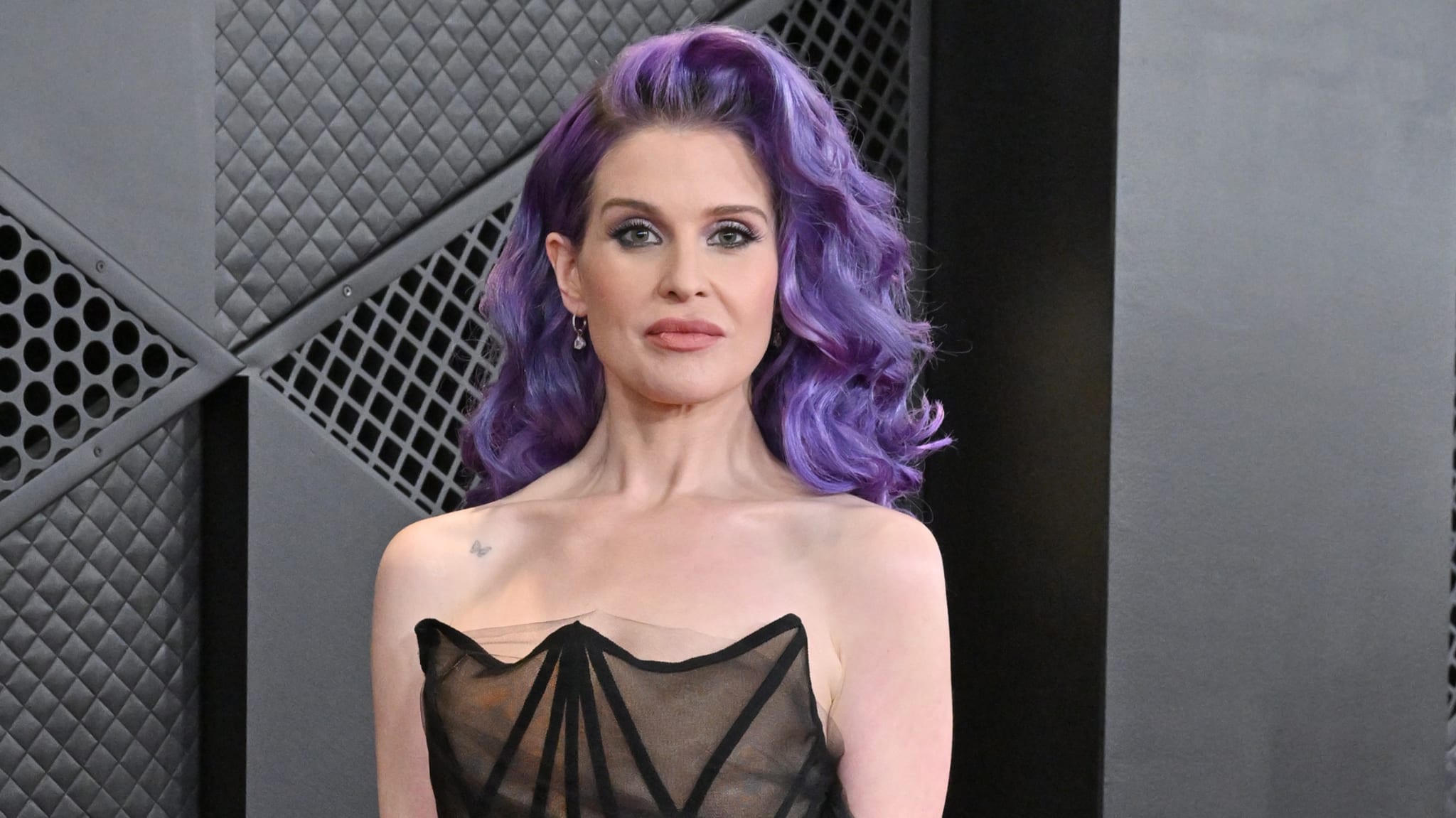 Woman with purple hair in black strap dress stands against patterned backdrop