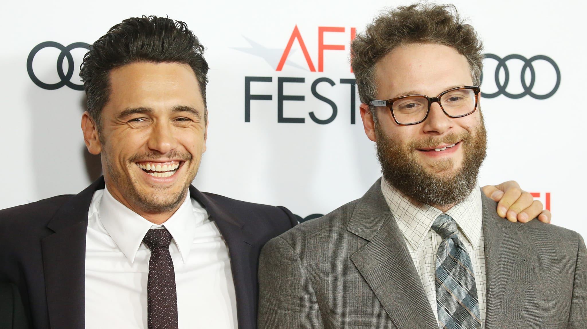 James Franco and Seth Rogen smiling at an AFI Fest event. Franco is in a dark suit, and Rogen wears glasses and a plaid tie.