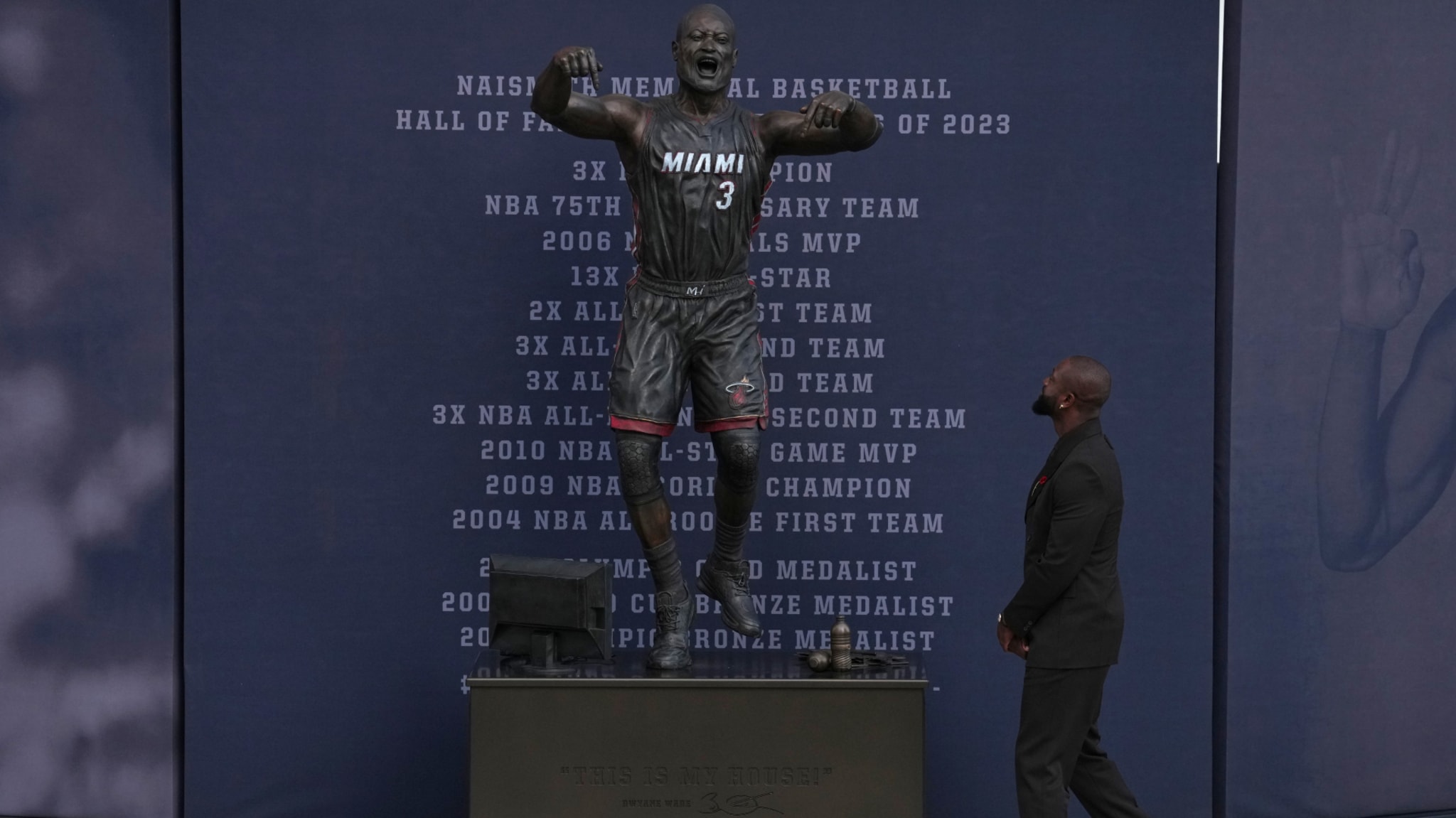 A man in a suit looks at a statue of a basketball player in a Miami jersey, with a backdrop listing achievements.