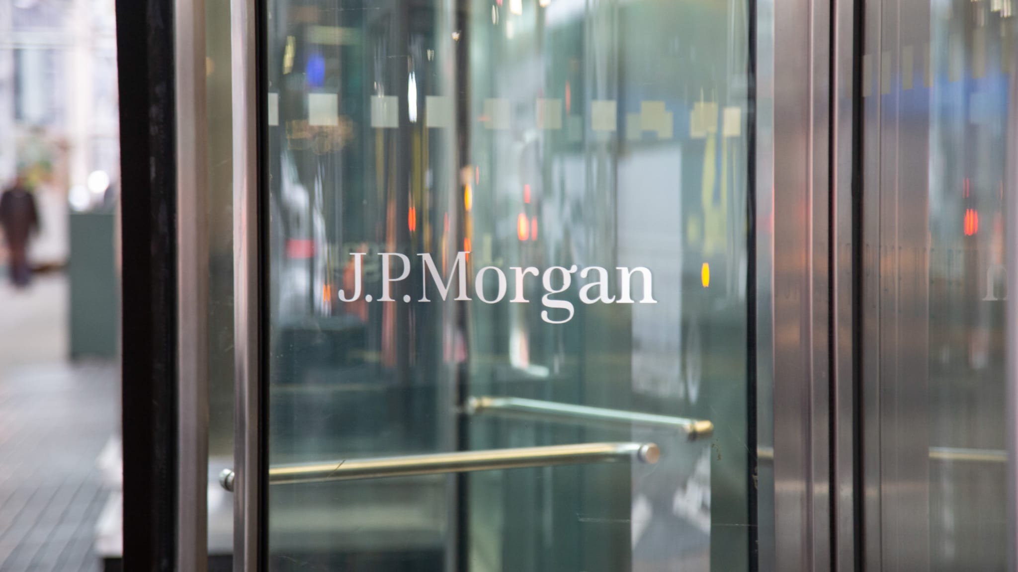 Glass door with "J.P. Morgan" logo in white lettering, reflecting urban street scene.