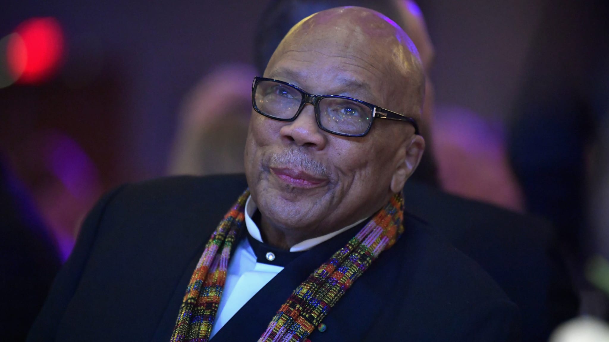 Quincy Jones, wearing glasses and a colorful scarf, is seated, smiling slightly. The background is softly illuminated.