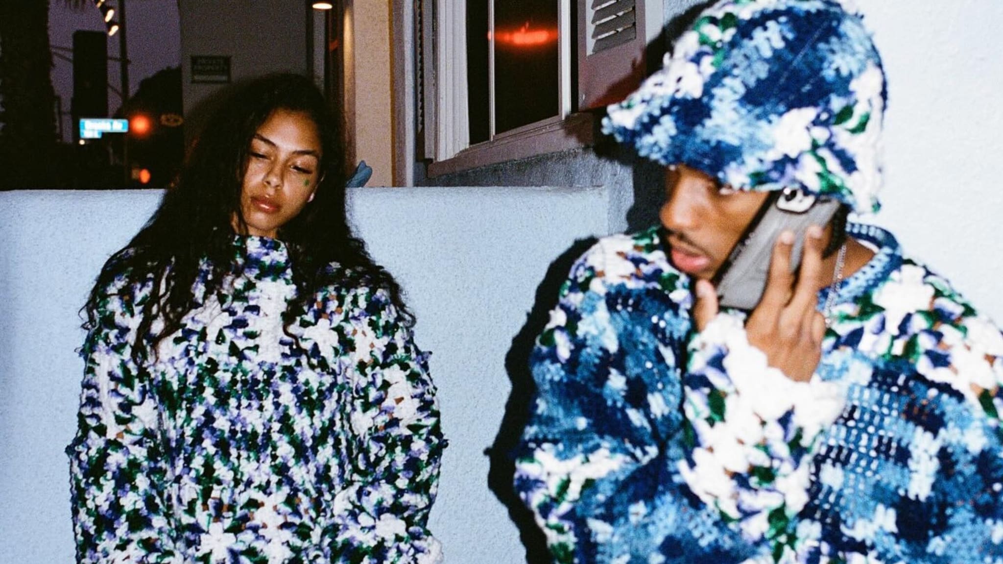 Two people wearing matching blue and white crocheted outfits. One is on the phone, and the other leans against a wall. Nigel Xavier