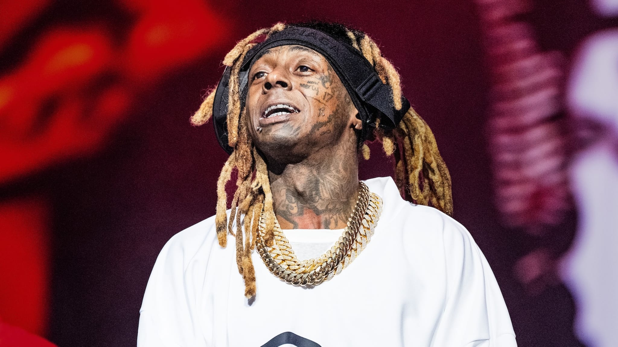Lil Wayne performing on stage, wearing a white shirt, gold chains, and a headband with his signature dreadlocks.