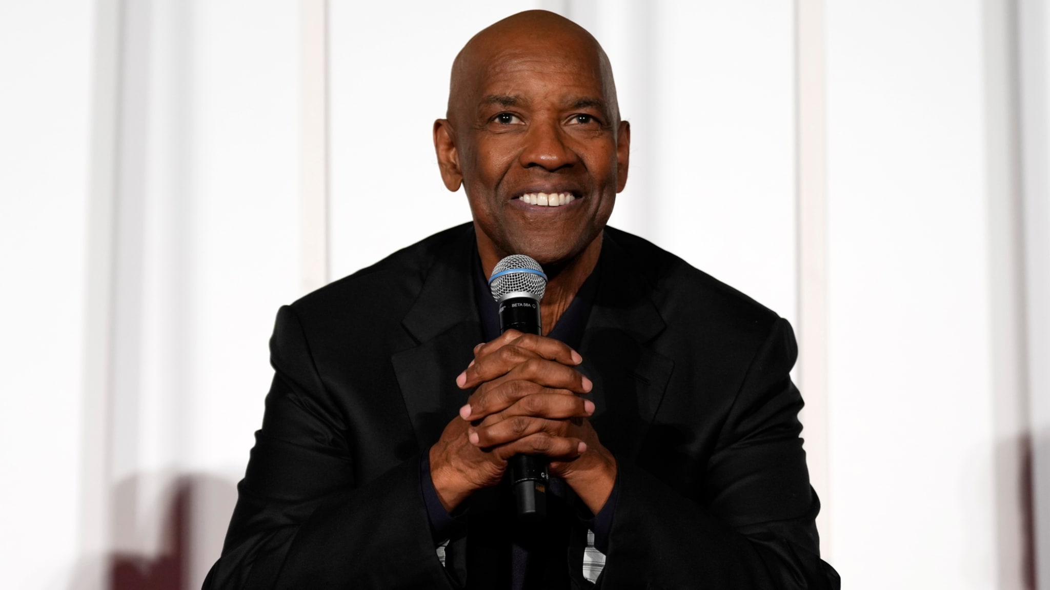 Denzel Washington, wearing a black suit, smiles while holding a microphone.