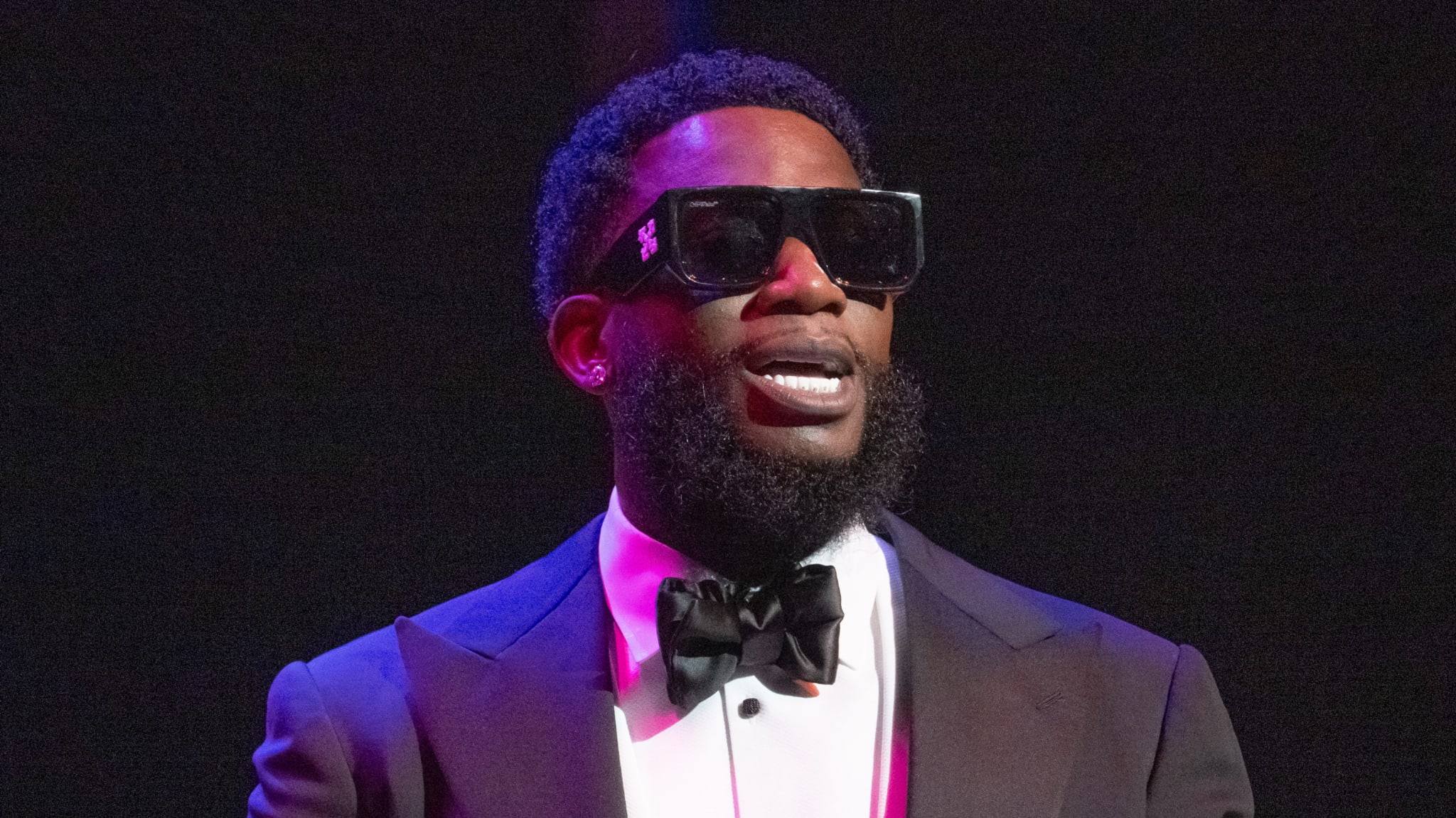 A man in a tuxedo and large sunglasses, with a beard, stands against a dark background.