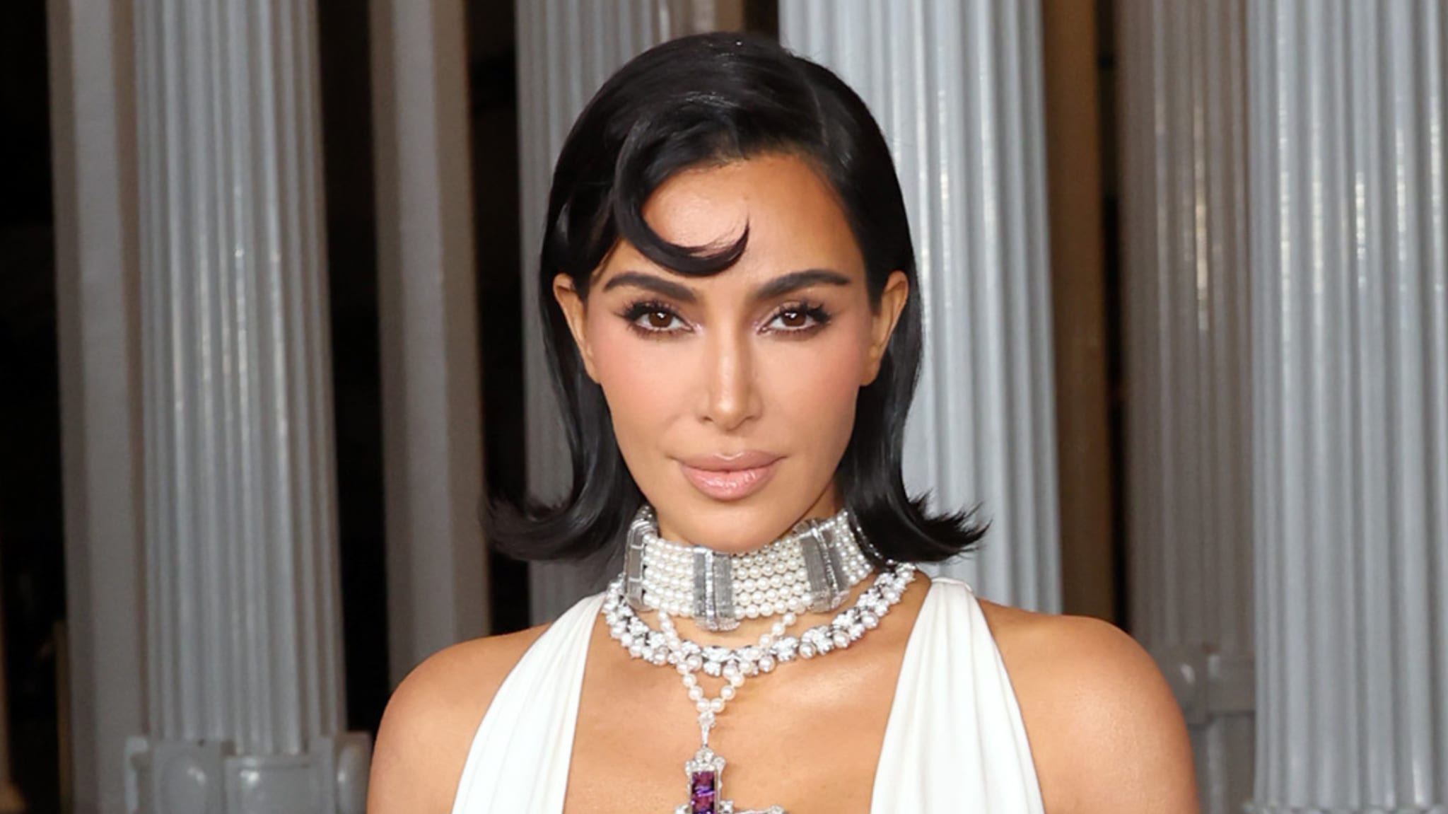 Kim Kardashian wearing a white dress and pearl necklace, with sleek short hair, standing in front of a columned backdrop.