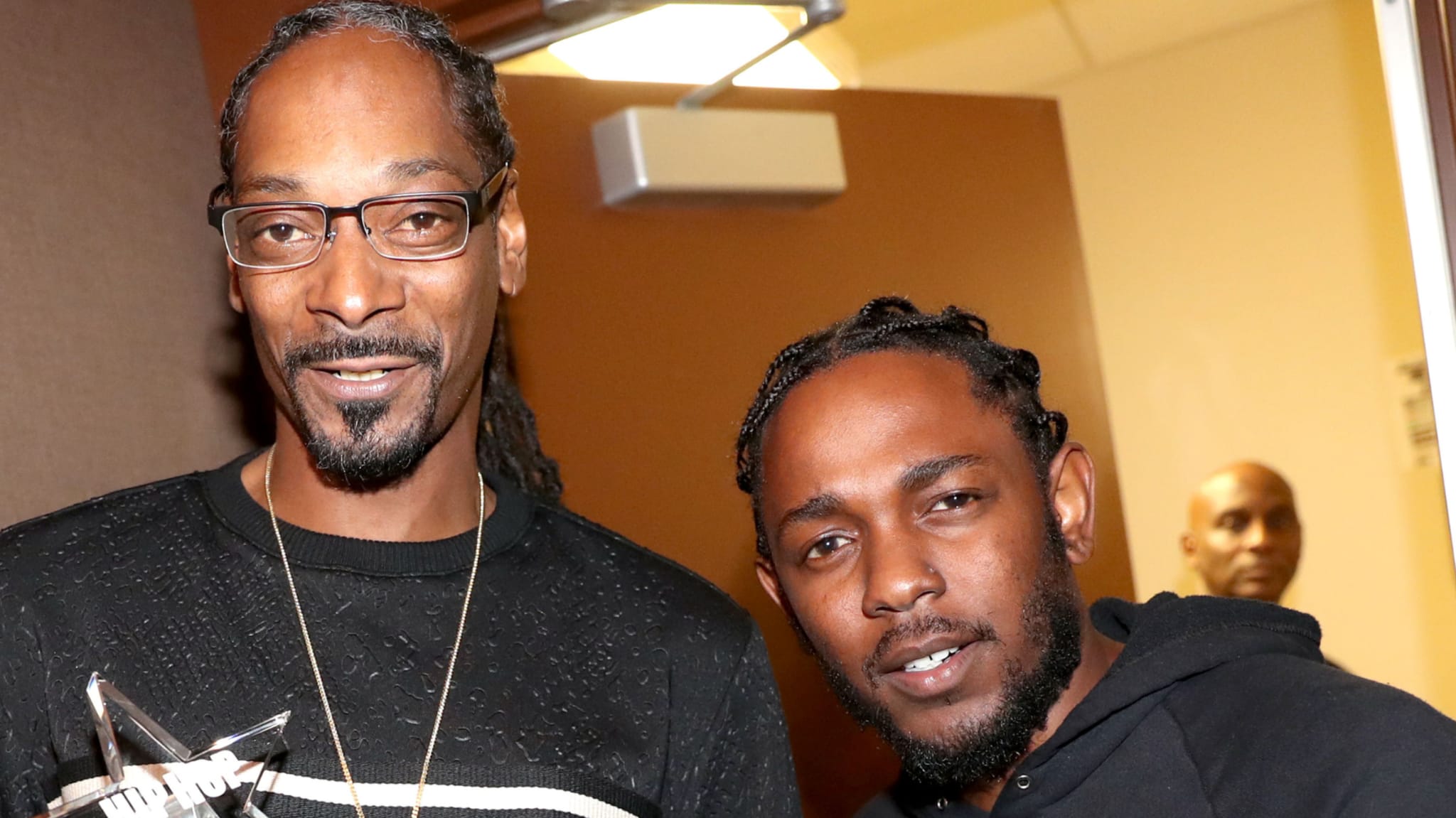 Snoop Dogg and Kendrick Lamar pose together, both smiling. Snoop wears glasses and a black sweater, while Kendrick wears a black hoodie.
