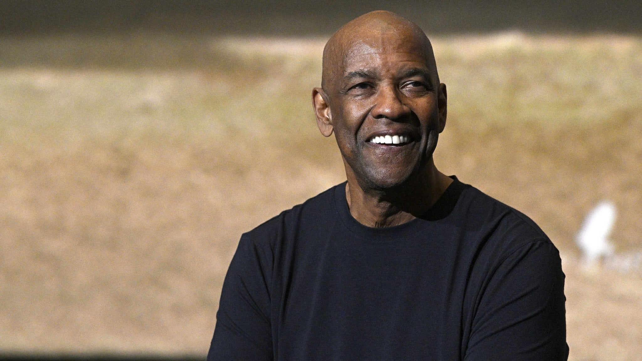 Denzel Washington smiling, wearing a black shirt, with a blurred background.