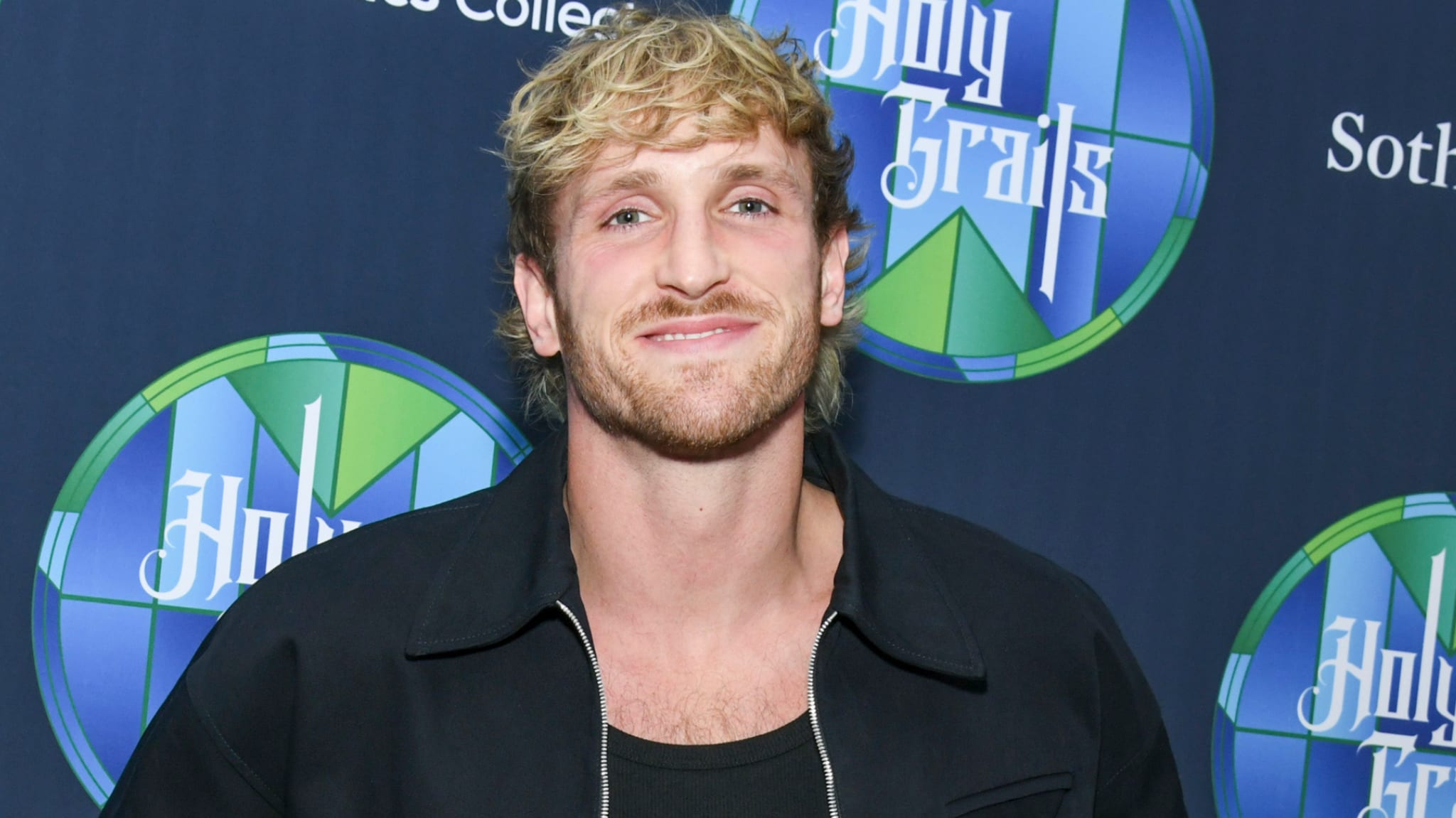Logan Paul smiling at an event, wearing a black jacket. A blue and green logo is in the background.