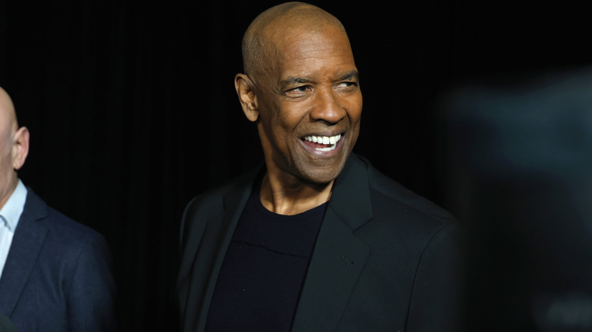 Denzel Washington smiling, wearing a black suit, against a dark background.