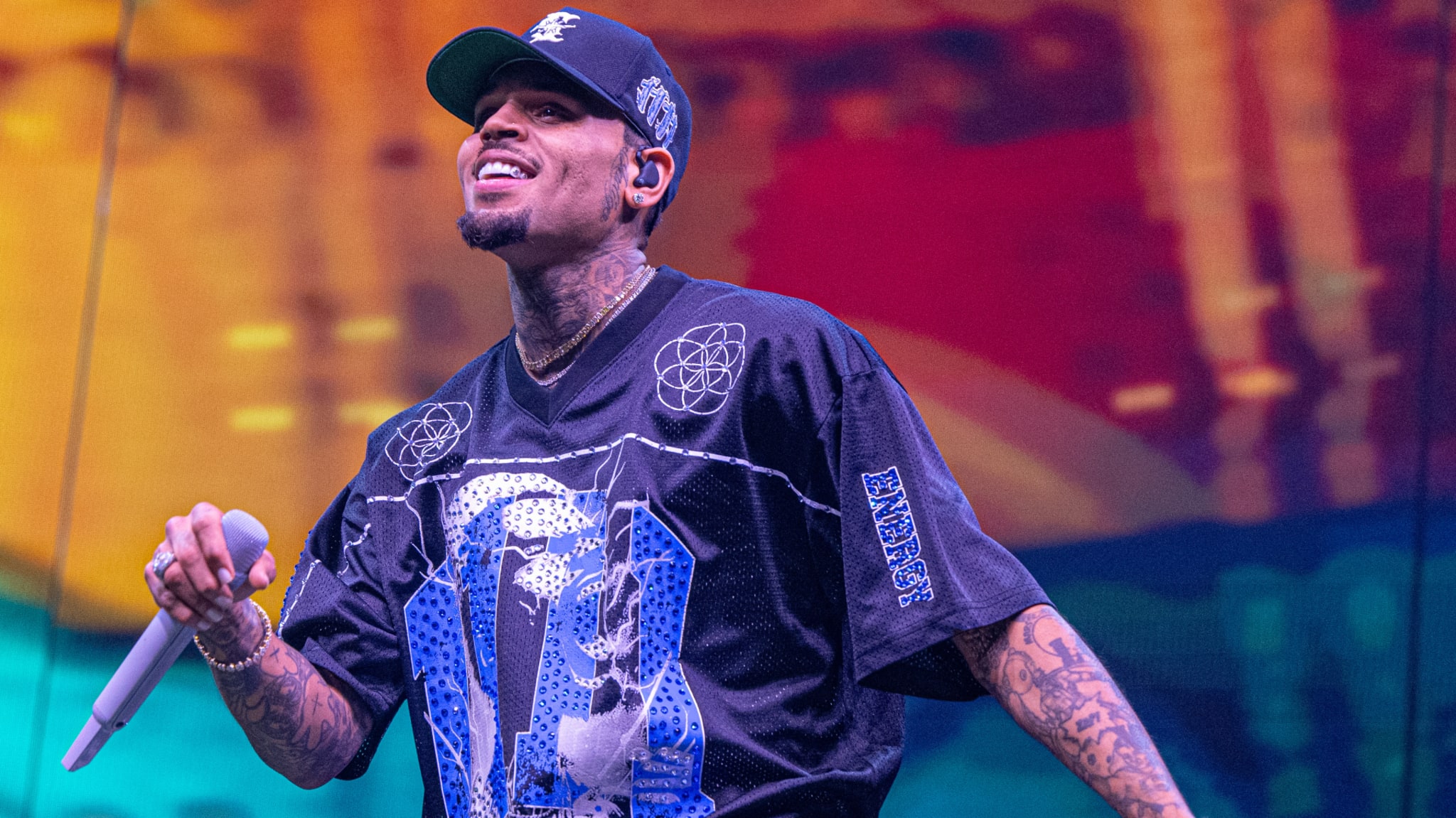 Chris Brown performing on stage, wearing a black graphic shirt and cap, holding a microphone, with a colorful background.