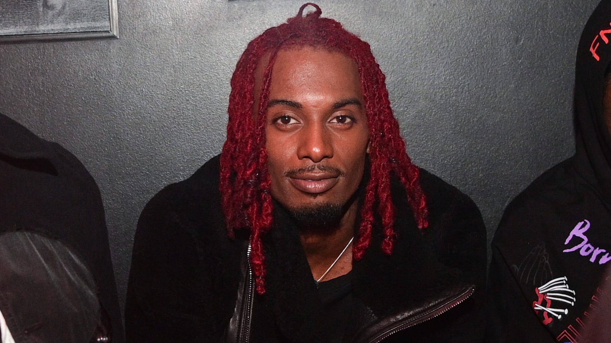A man with red dreadlocks and a black jacket sits against a dark background.