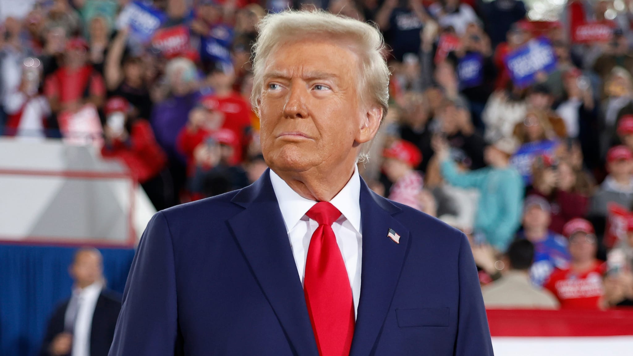 Donald Trump at a rally, wearing a navy suit and red tie, with a crowd of supporters in the background.
