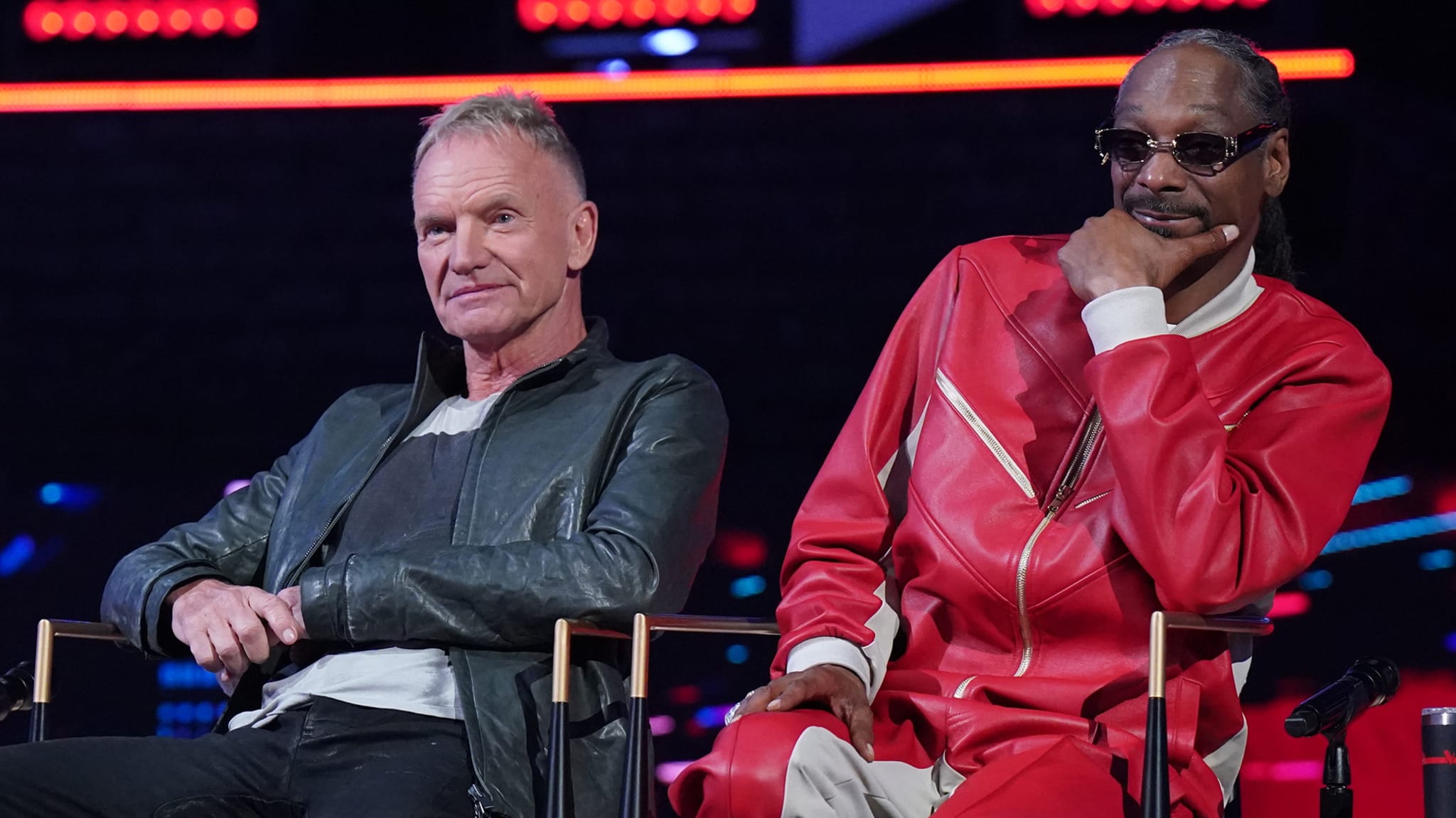 Sting and Snoop Dogg sit together. Sting wears a dark jacket, while Snoop Dogg sports a red and white outfit with sunglasses.
