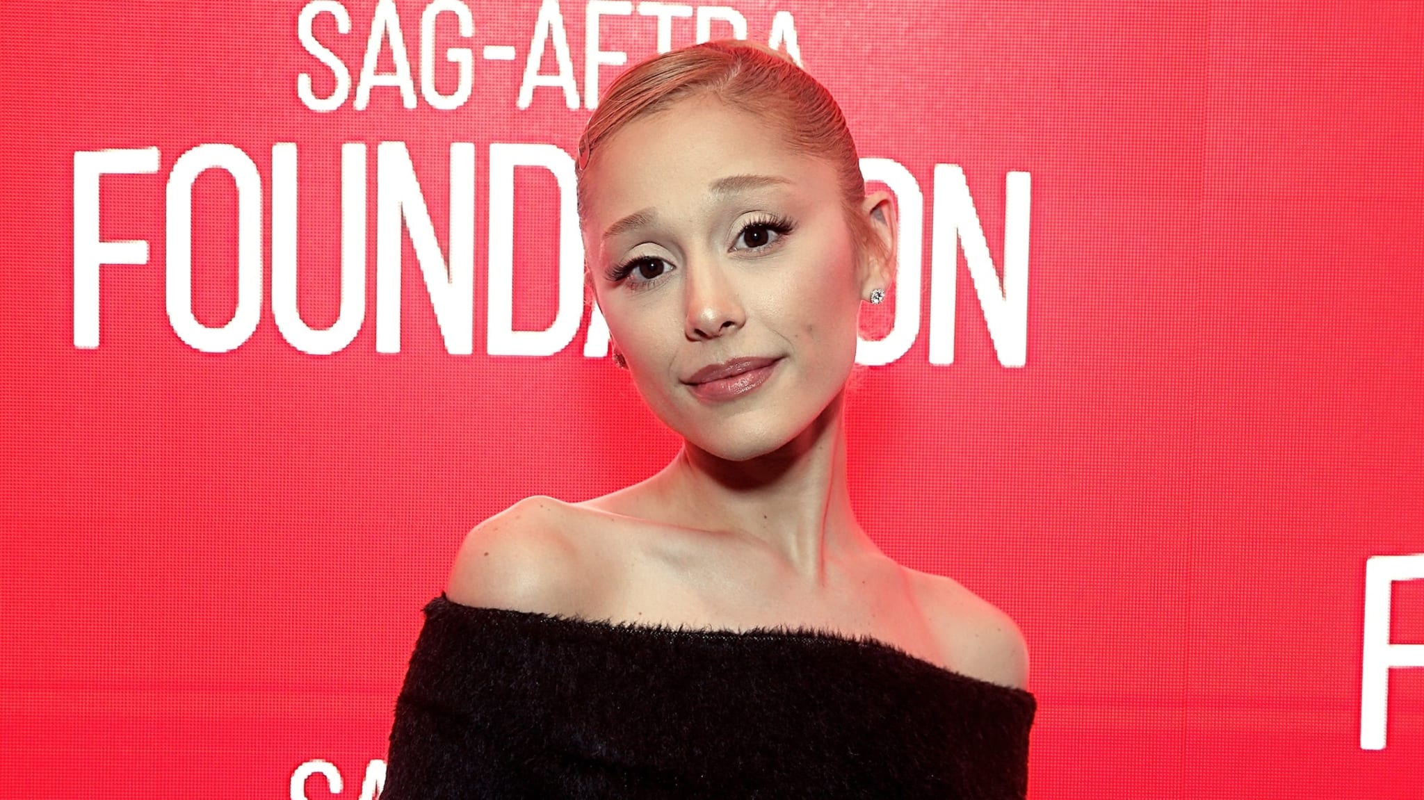 Ariana Grande with a sleek bun and off-shoulder black dress stands in front of a red background with "SAG-AFTRA Foundation" text.