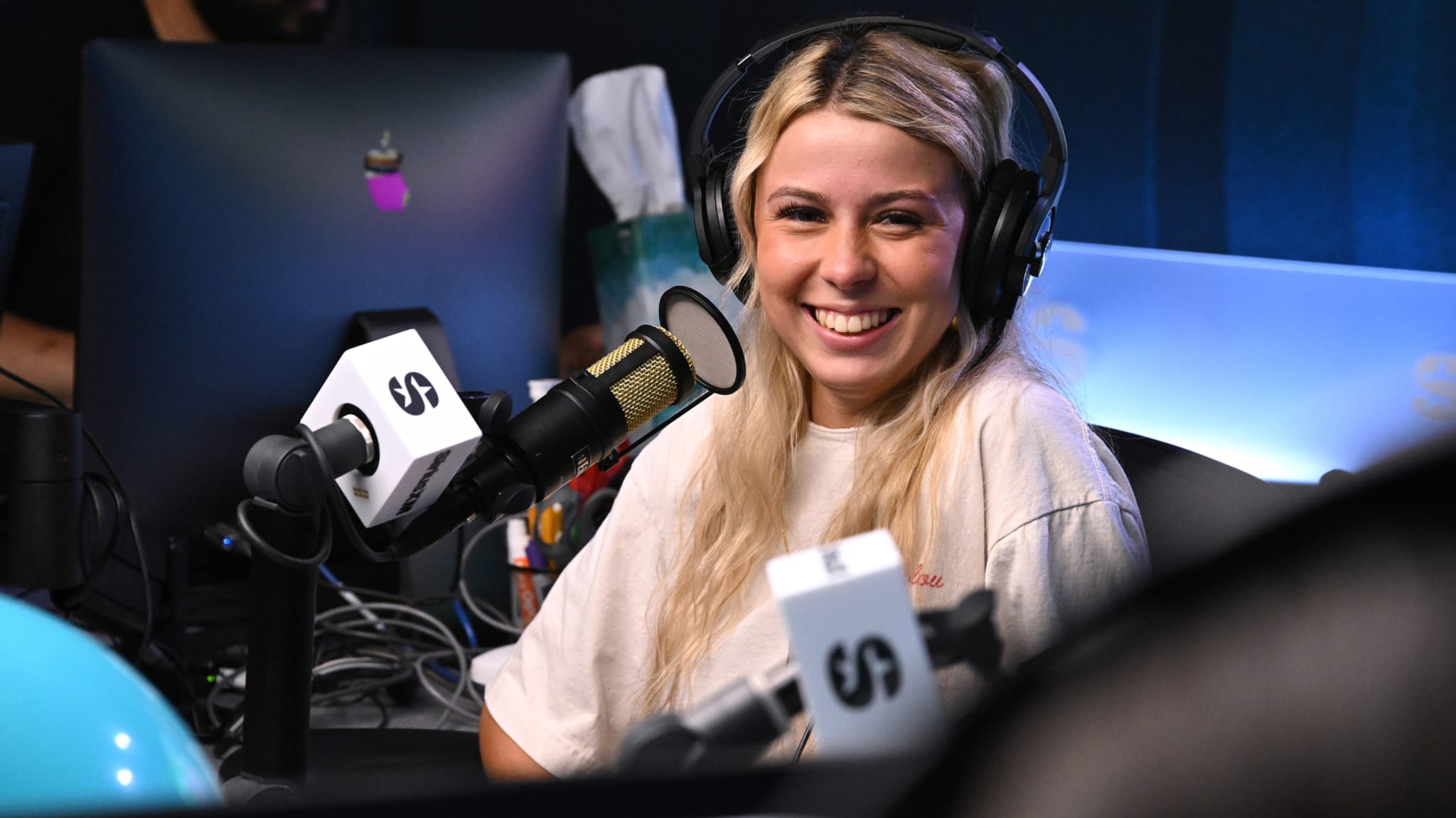 A woman with blonde hair is smiling while wearing headphones and speaking into a microphone in a radio studio.