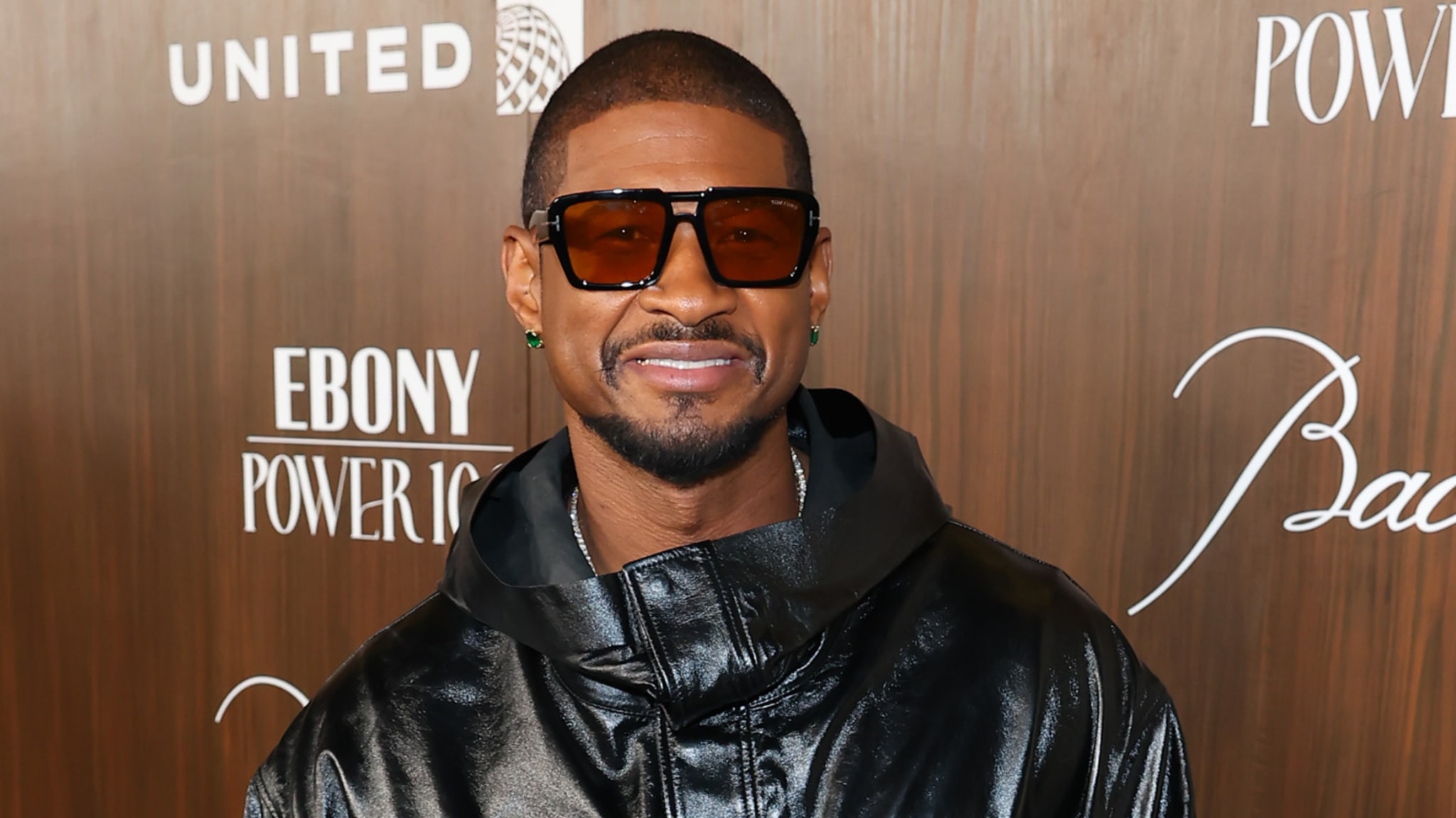 Usher wearing sunglasses and a black leather jacket at an event, smiling in front of a wooden backdrop with logos.
