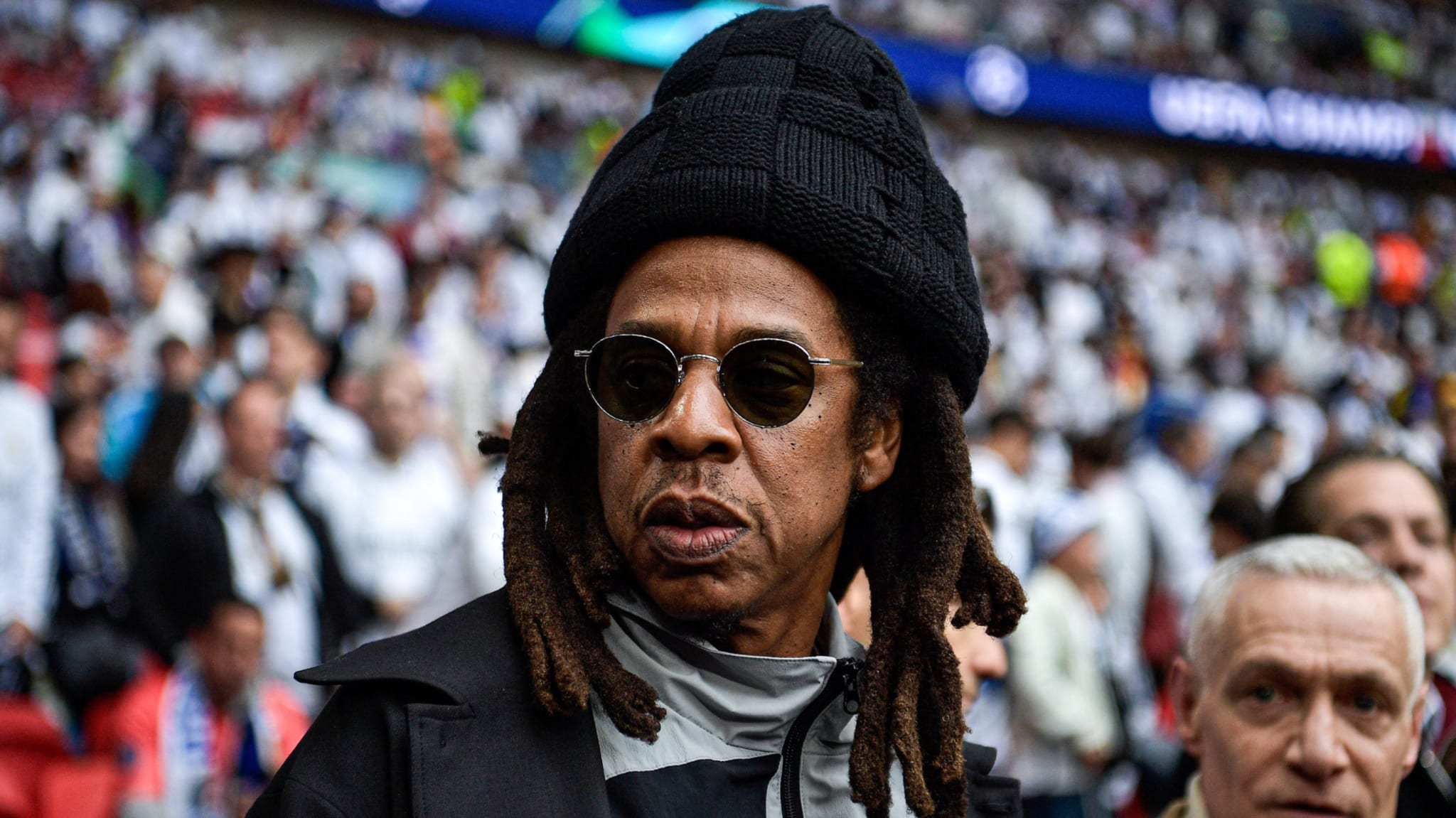 A man with dreadlocks and sunglasses wearing a black hat and coat, standing in a crowded stadium.
