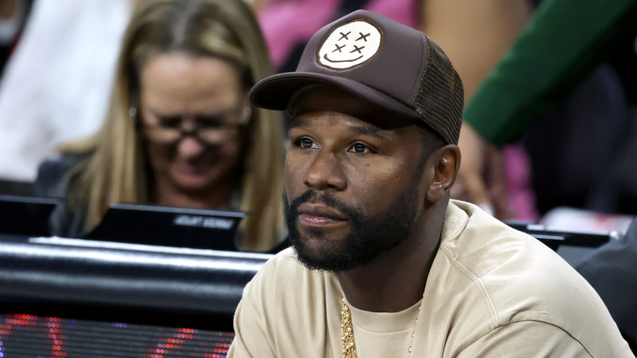 Floyd Mayweather, wearing a brown cap and beige shirt, sits at an event.