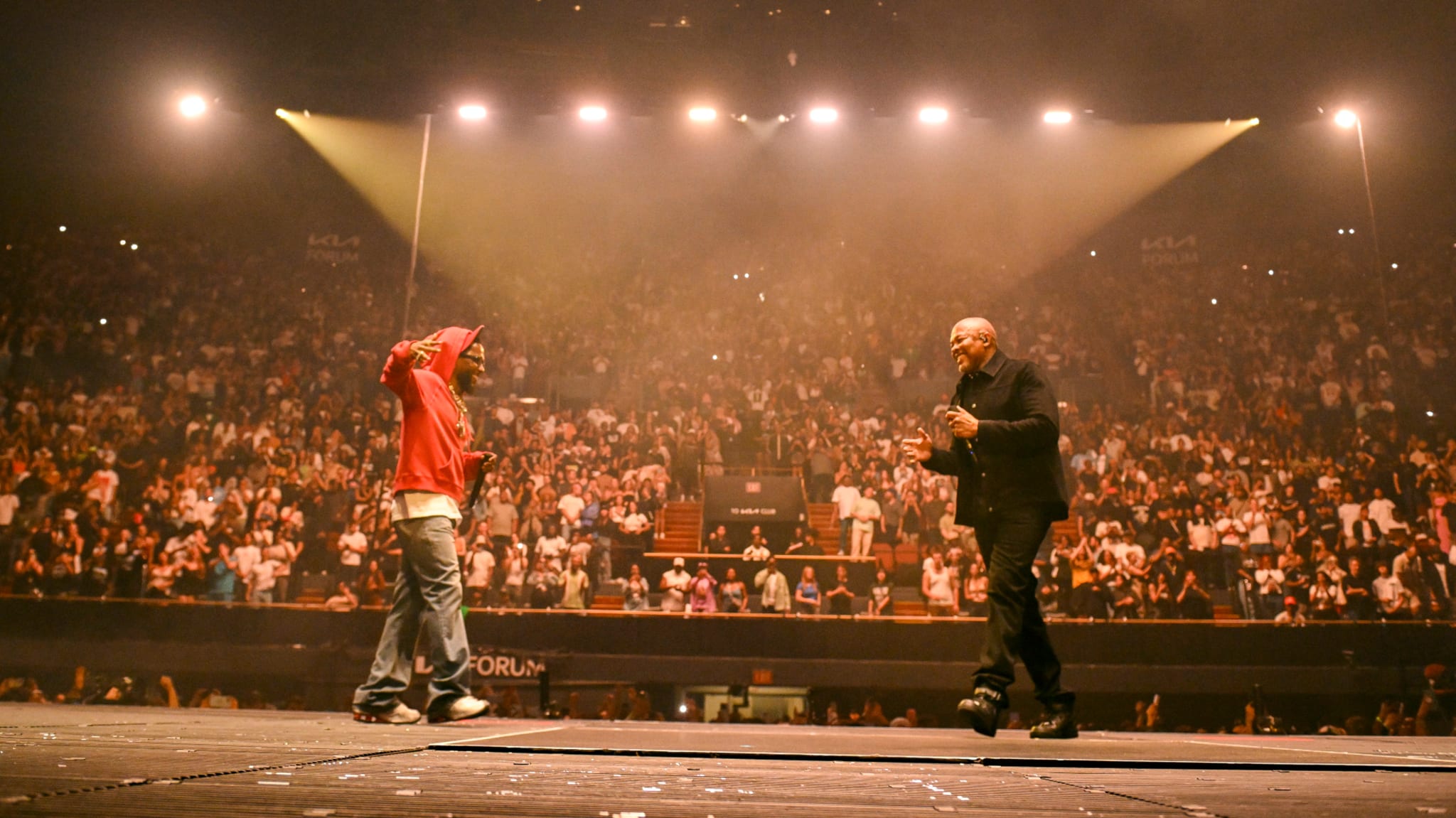 Two performers on stage in front of a large audience. One wears a red hoodie, the other in black. Bright lights illuminate the scene.