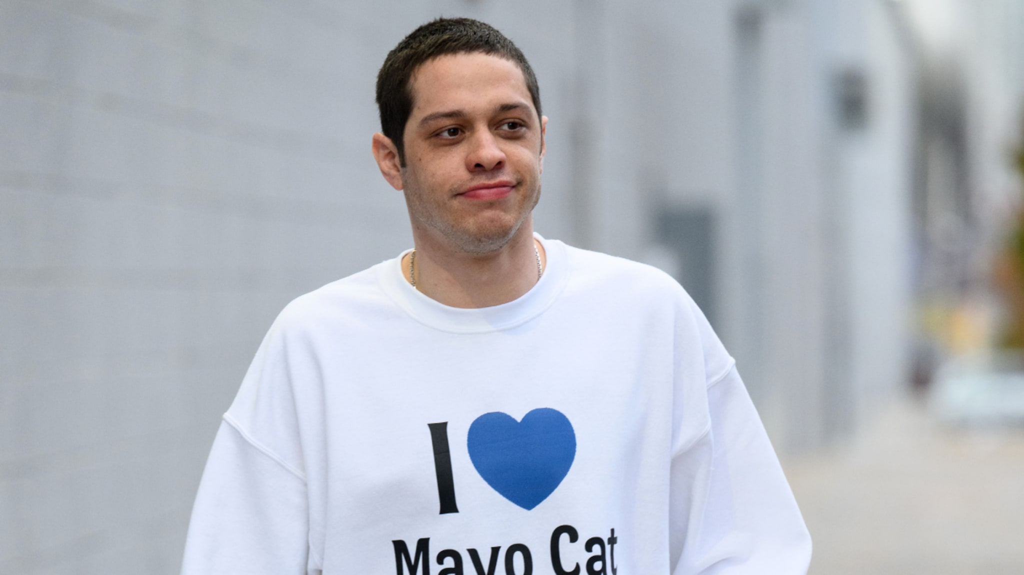 Pete Davidson wearing a white sweatshirt with "I [heart] Mayo Cat" printed on it, walking outdoors.