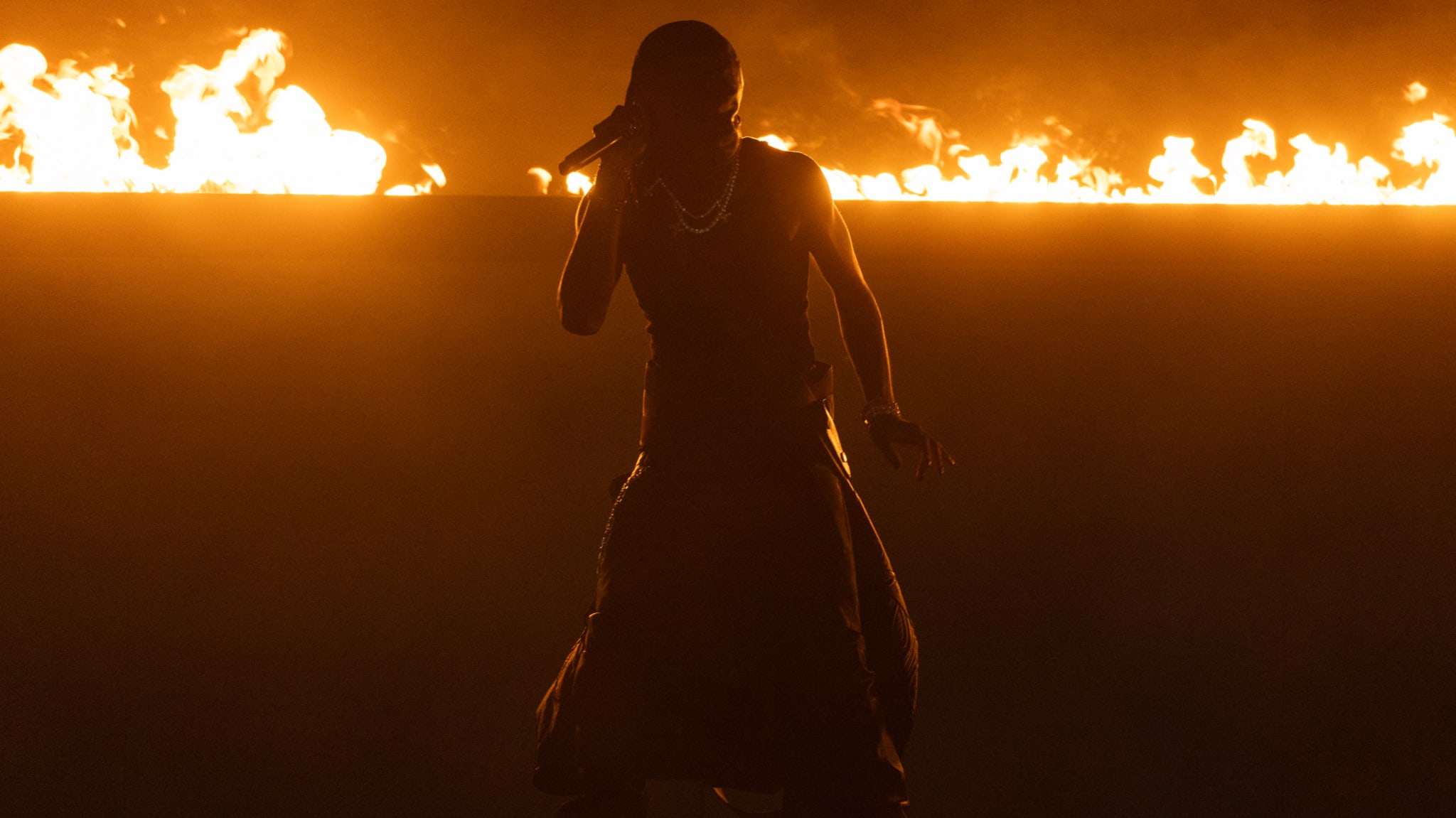 Silhouette of a performer on stage with fire in the background, holding a microphone.