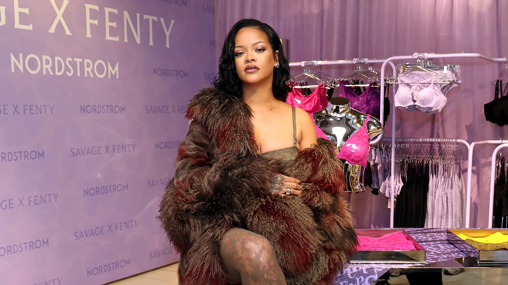 Rihanna at a Savage X Fenty event, wearing a fur coat. Lingerie displayed in the background with brand signage.