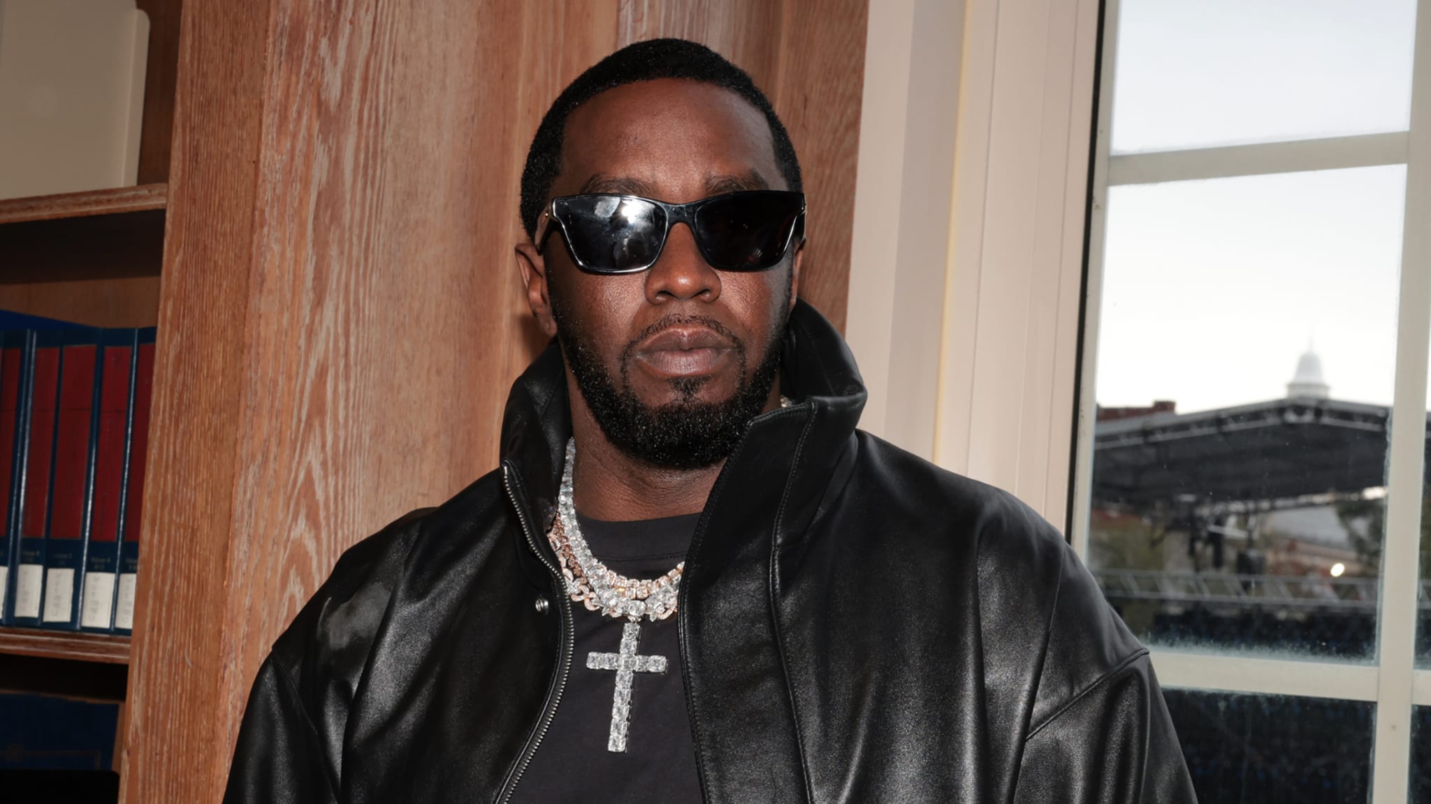 Sean "Diddy" Combs wearing sunglasses, a black leather jacket, and a diamond necklace, standing indoors.