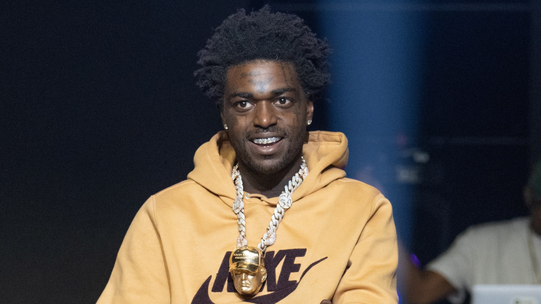 Rapper Kodak Black wearing a tan Nike hoodie and a large chain with a pendant, smiling on stage.