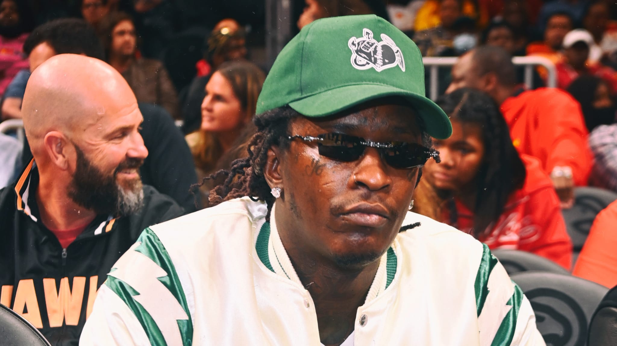 Young Thug wearing a green cap and sunglasses, sitting at an event.