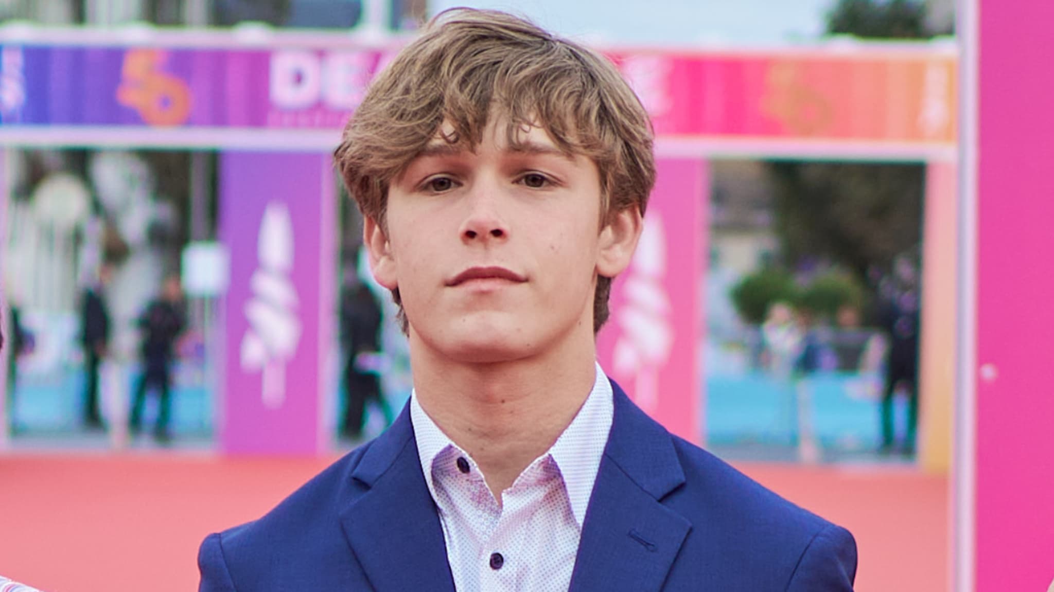 Hudson Meek in a blue suit on a red carpet, colorful background with blurred text and logos.