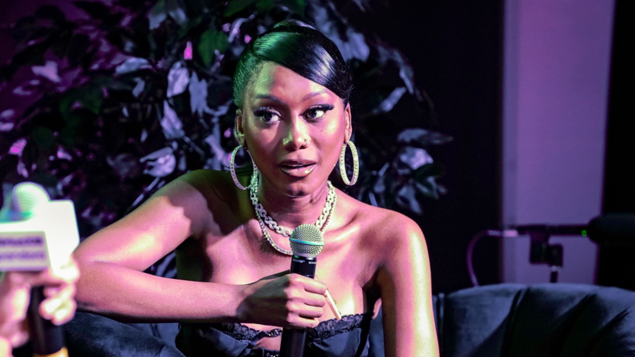 Muni Long with hoop earrings and a necklace holds a microphone, sitting on a stage with a plant in the background.