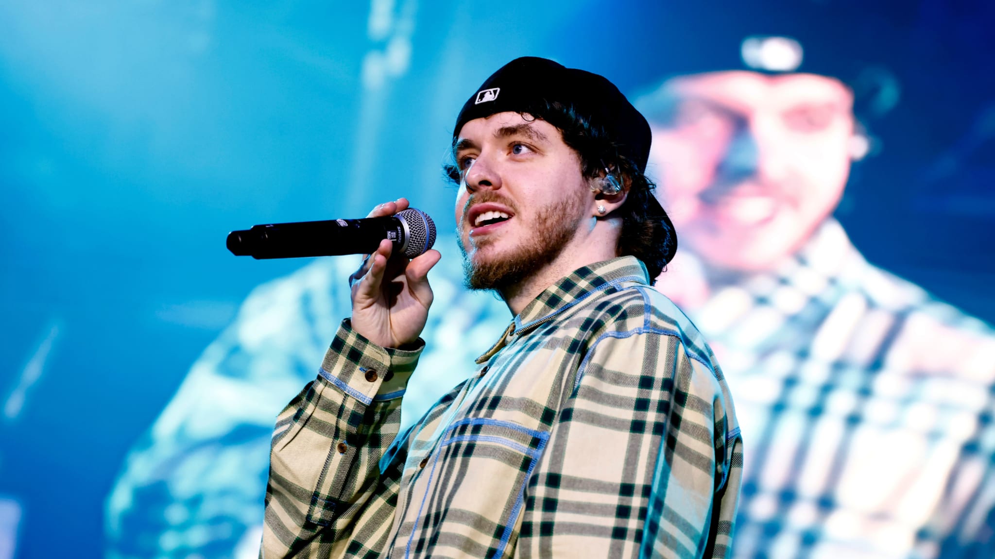 Jack Harlow performing on stage, wearing a plaid shirt and a black cap, holding a microphone with a large screen behind him.