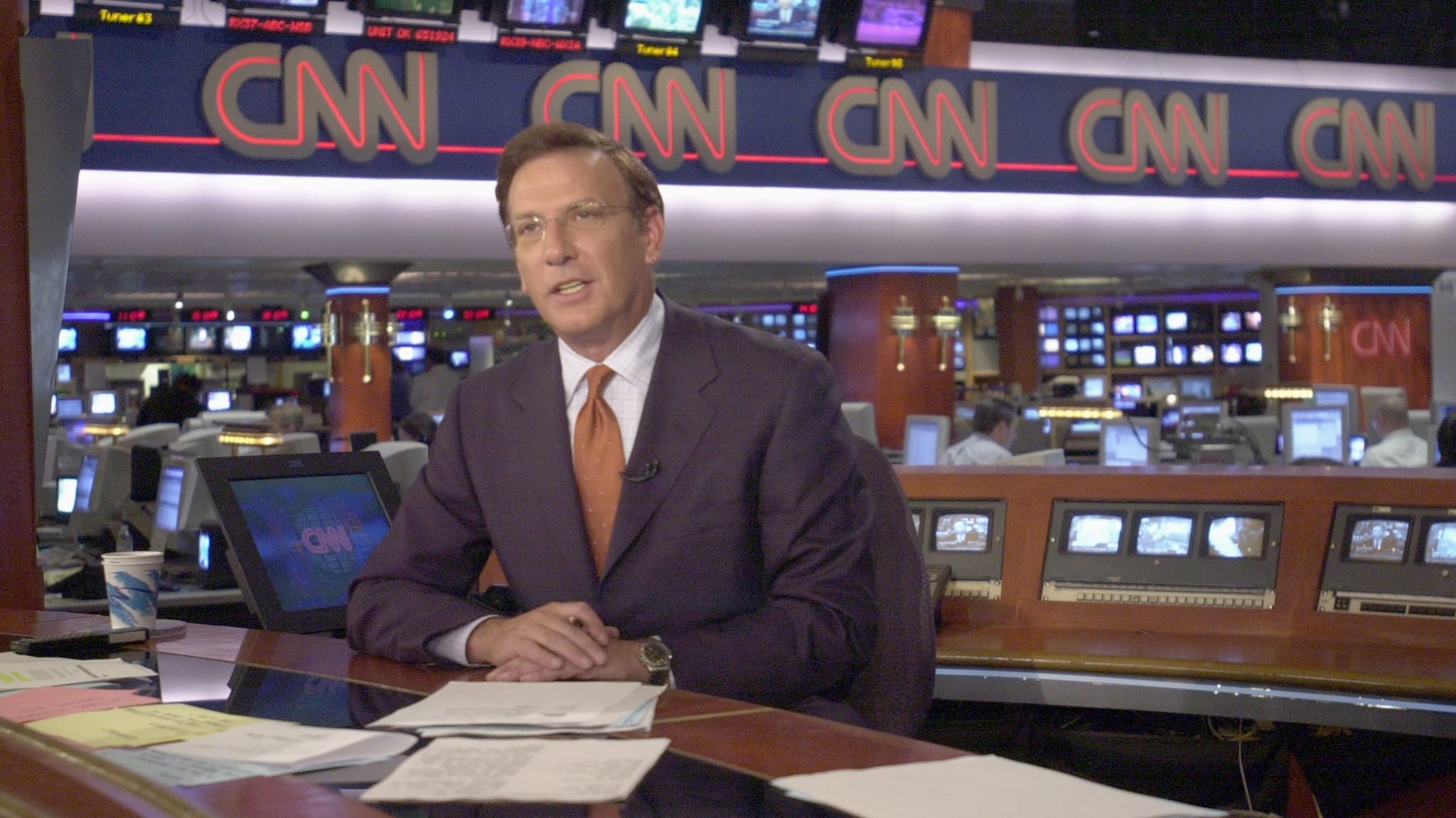 Aaron Brown in a suit and tie sits at a desk in a CNN newsroom, surrounded by monitors and papers.