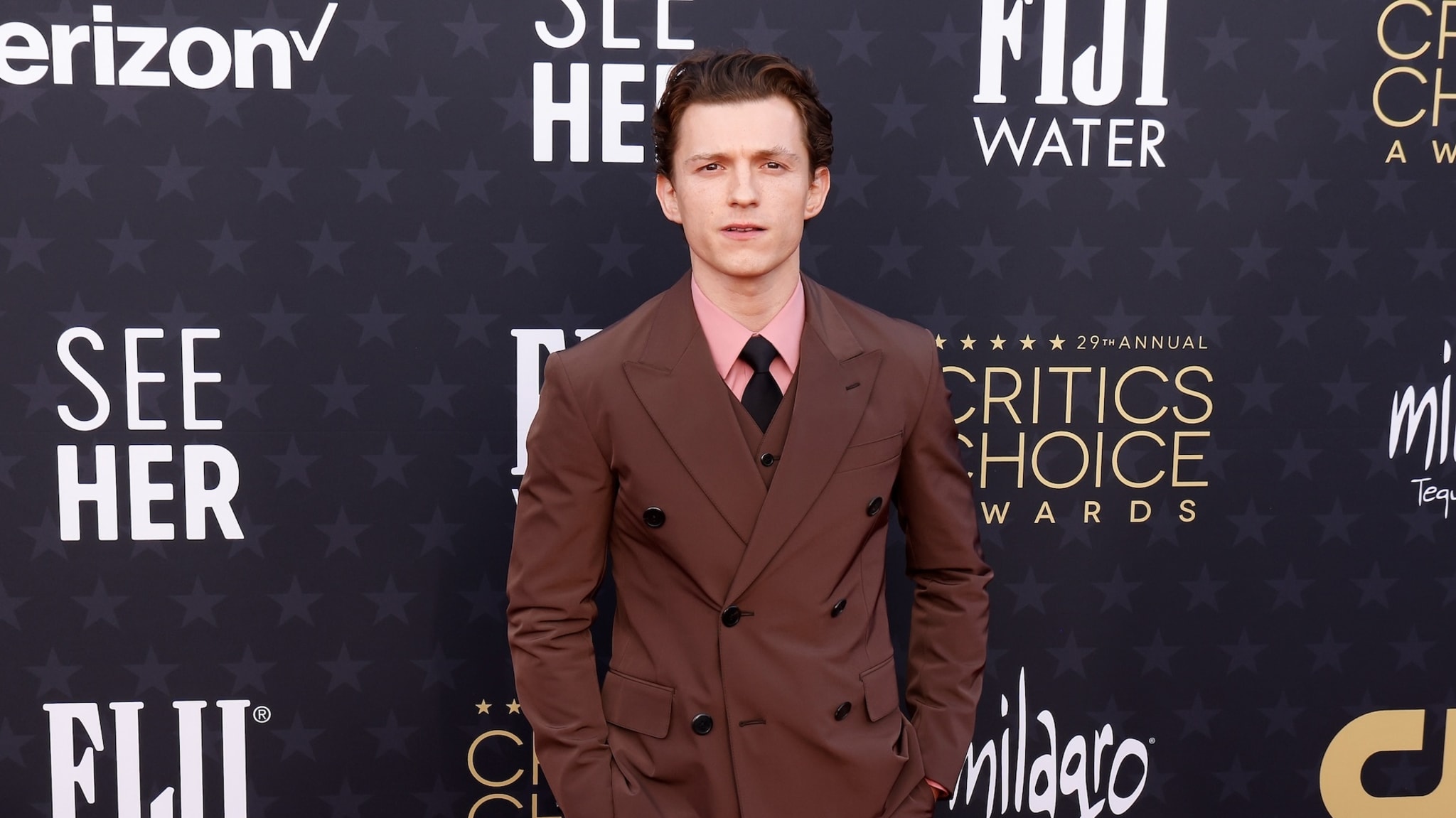 Tom Holland at the Critics Choice Awards, wearing a brown suit with a pink shirt and black tie, standing on the red carpet.