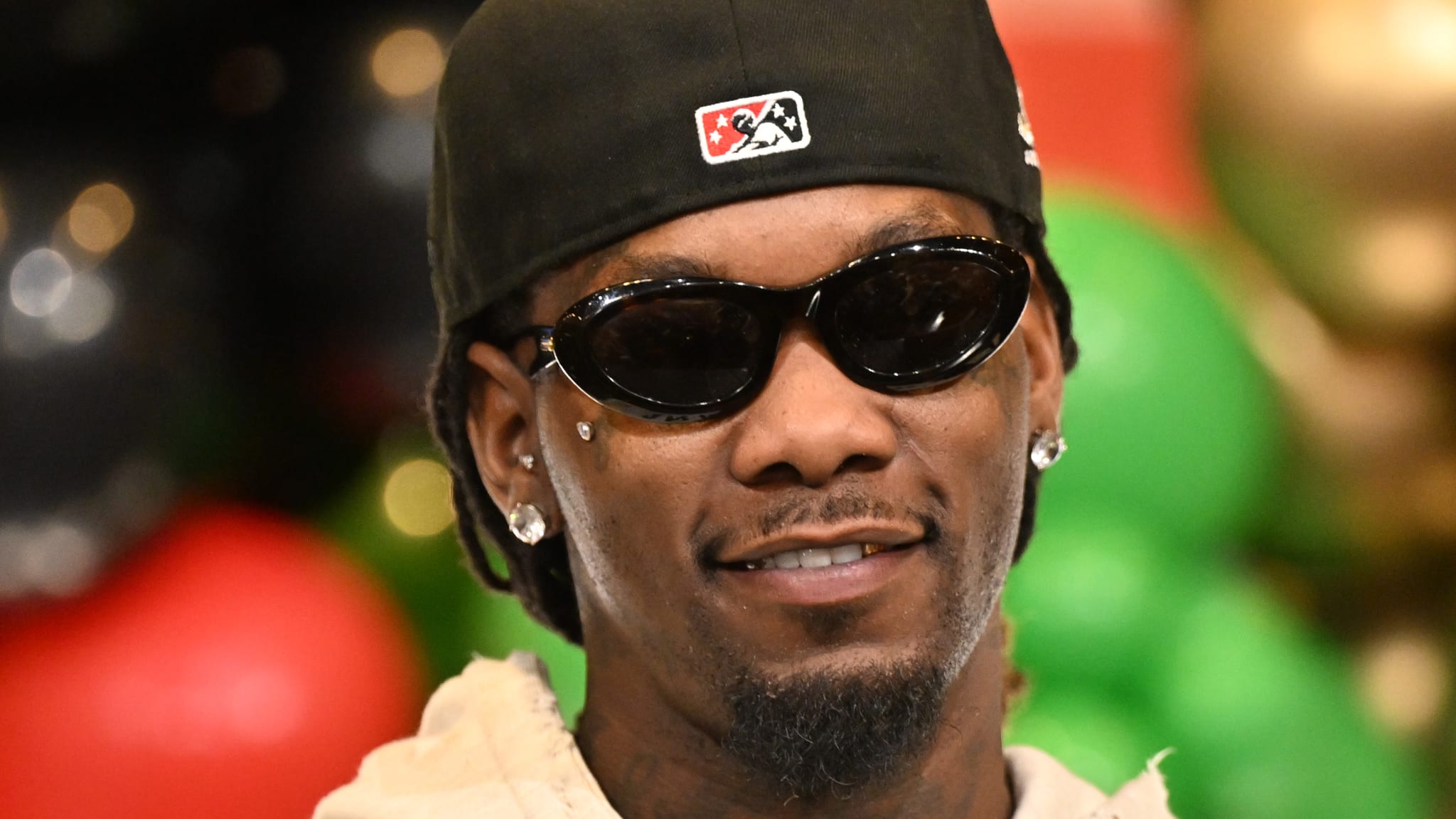 Offset wearing sunglasses and a cap, standing in front of colorful balloons, smiling.