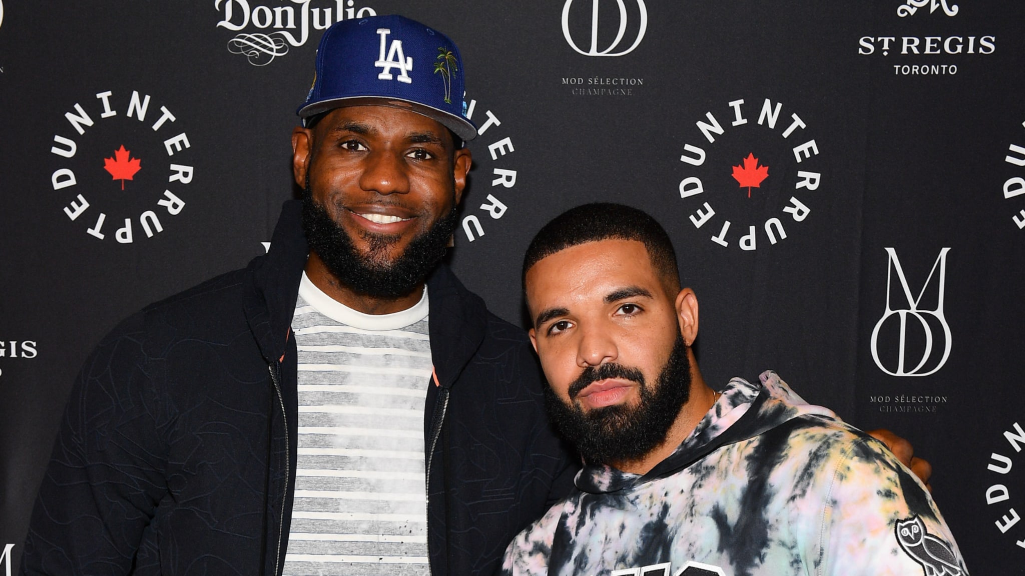 LeBron James wearing a blue LA cap stands with Drake in a tie-dye hoodie at an event with branded backdrops.